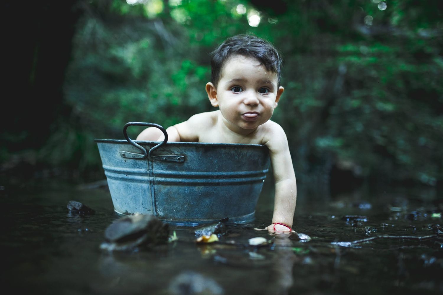A curious baby sits in a vintage metal washtub outdoors in a shallow woodland stream surrounded by rocks.