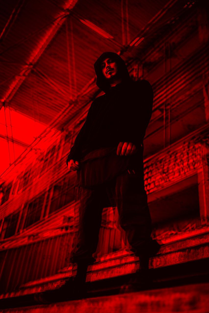 Mysterious figure in dark hoodie standing on structure bathed in dramatic red light from below.