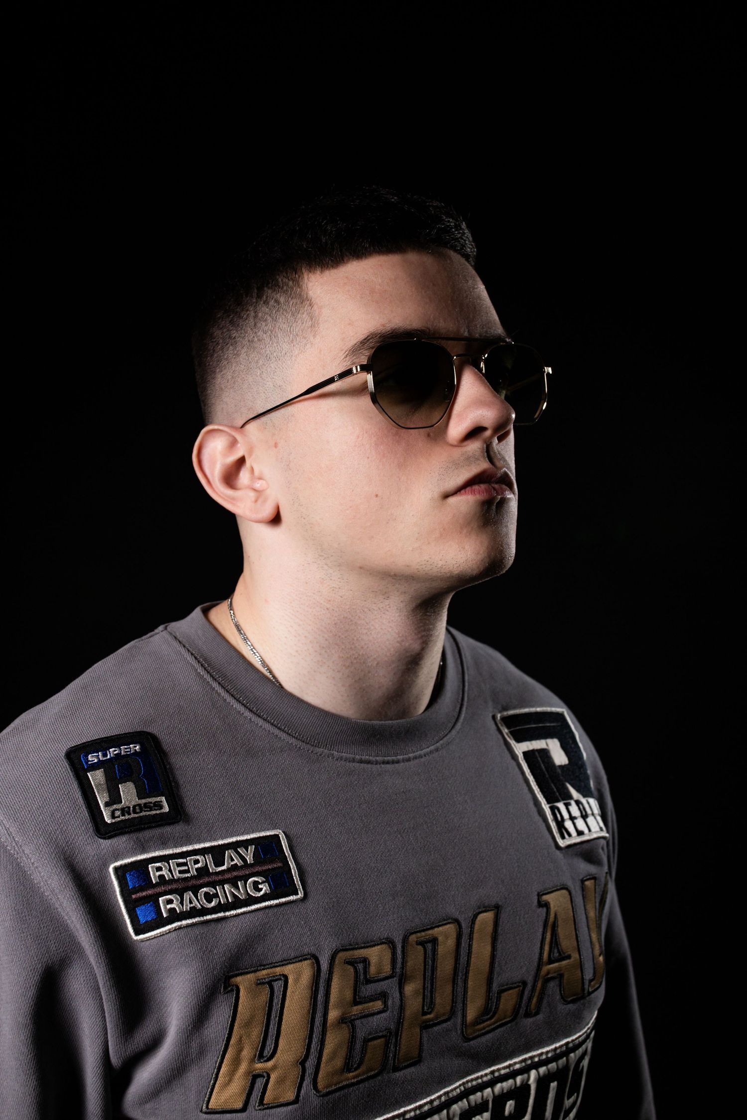 Young man in gray Replay Racing sweatshirt wearing aviator sunglasses against black background.
