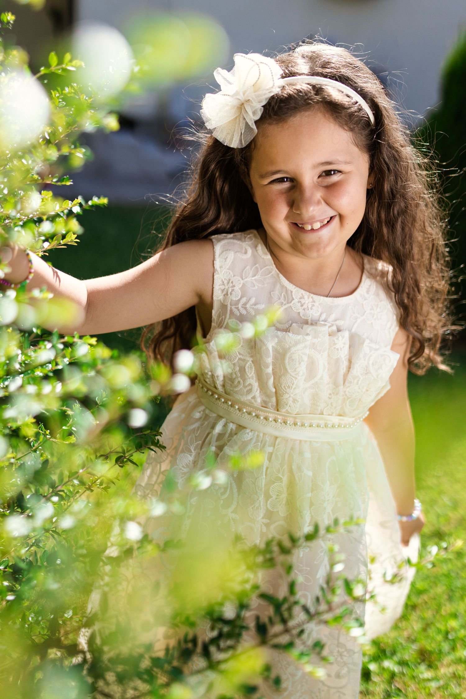 A smiling young girl in a white lace dress with floral headband stands among green bushes in a sunny garden.