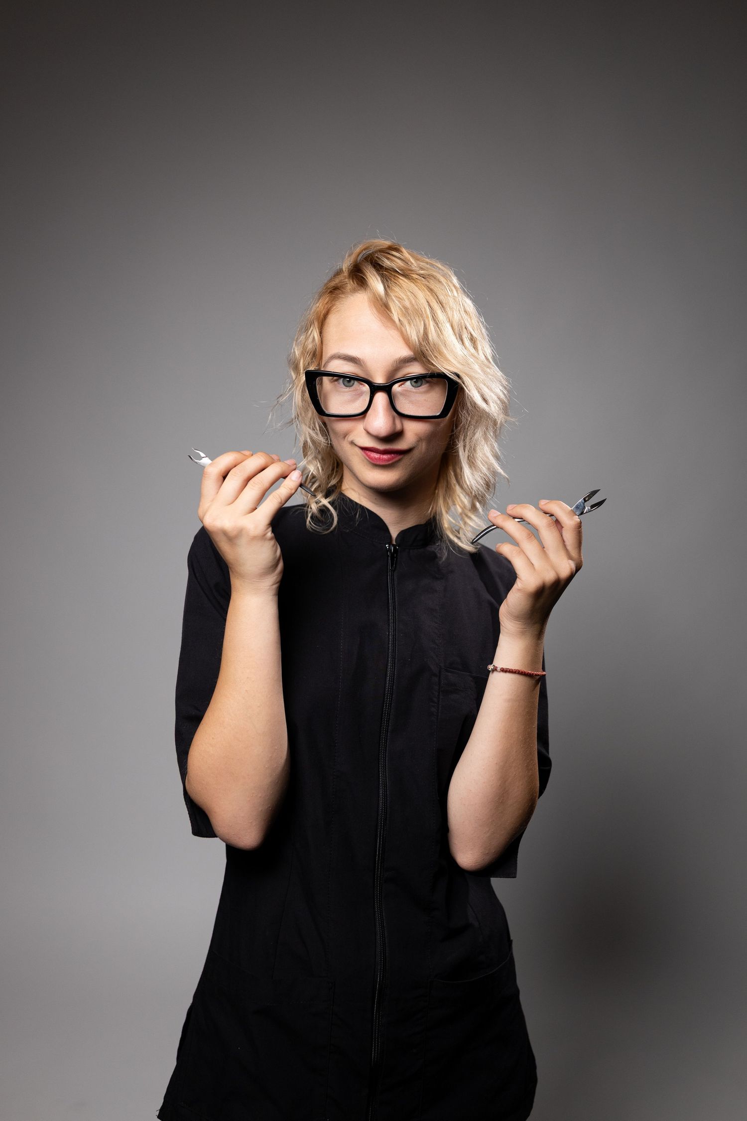 Blonde woman in black uniform and glasses holding small tools, smiling confidently against gray background.