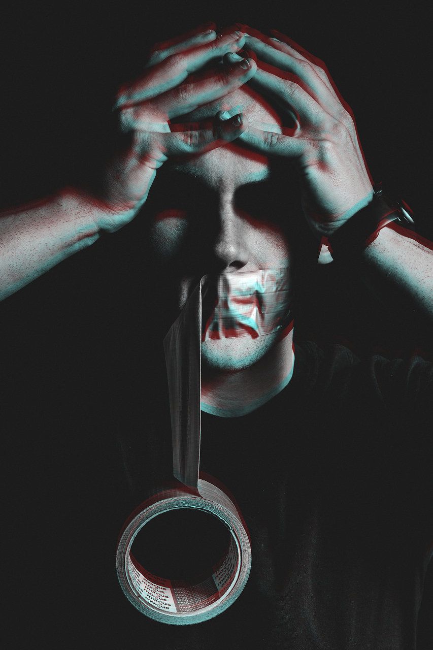 Man with tape over mouth holding head in hands with glitch effect on dark background.