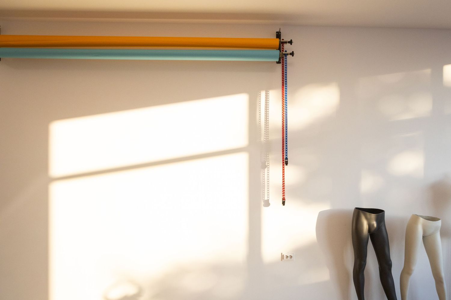 Sunlit white room with colorful wall-mounted rods and partial mannequin legs visible in corner.