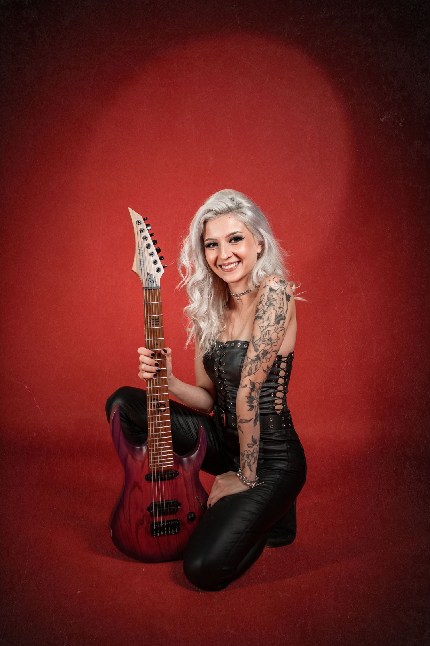 Smiling woman with silver hair and arm tattoos holding electric guitar wearing black corset on red background.