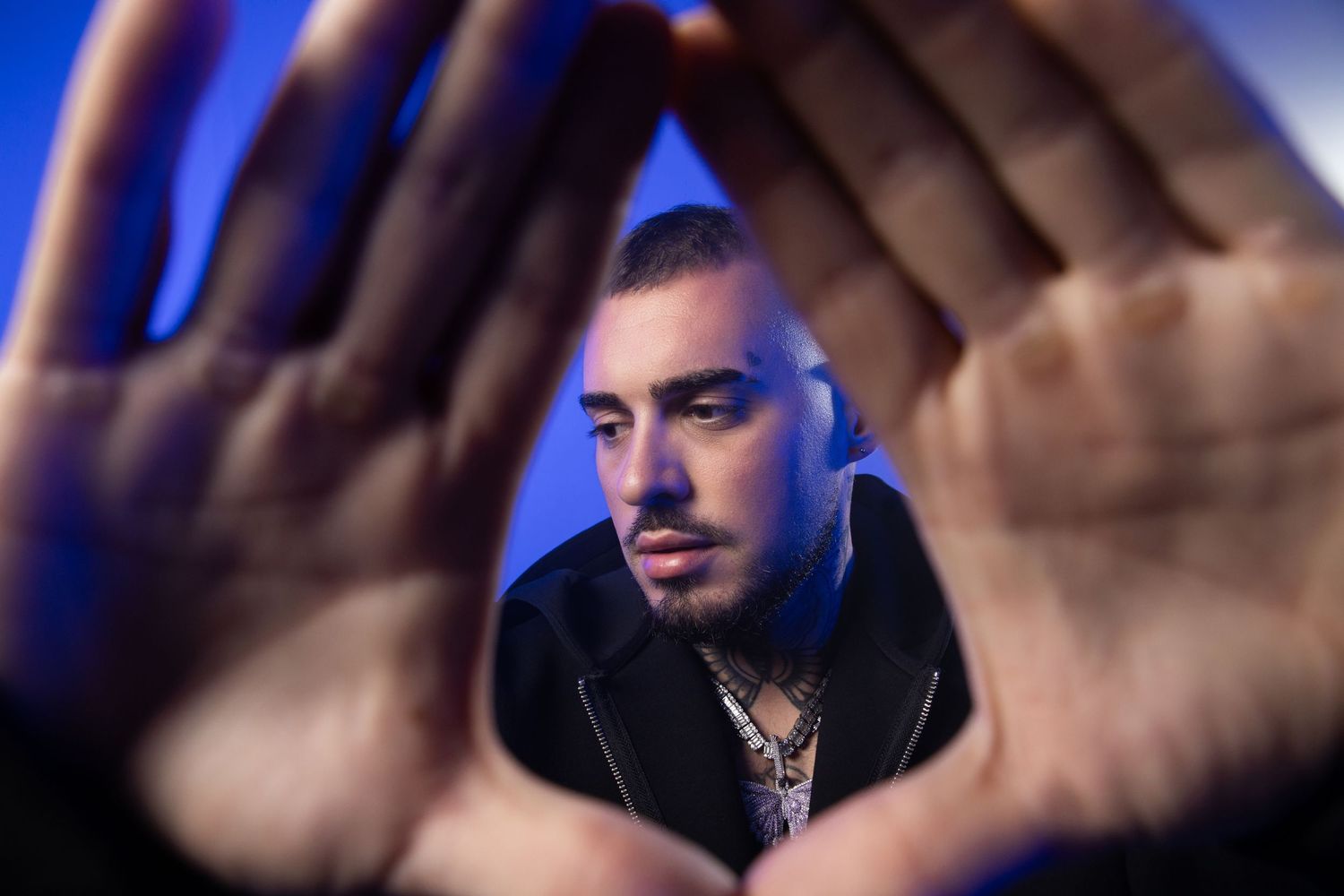Tattooed male artist framing face with hands in blue-lit studio, wearing black jacket with chain necklace.