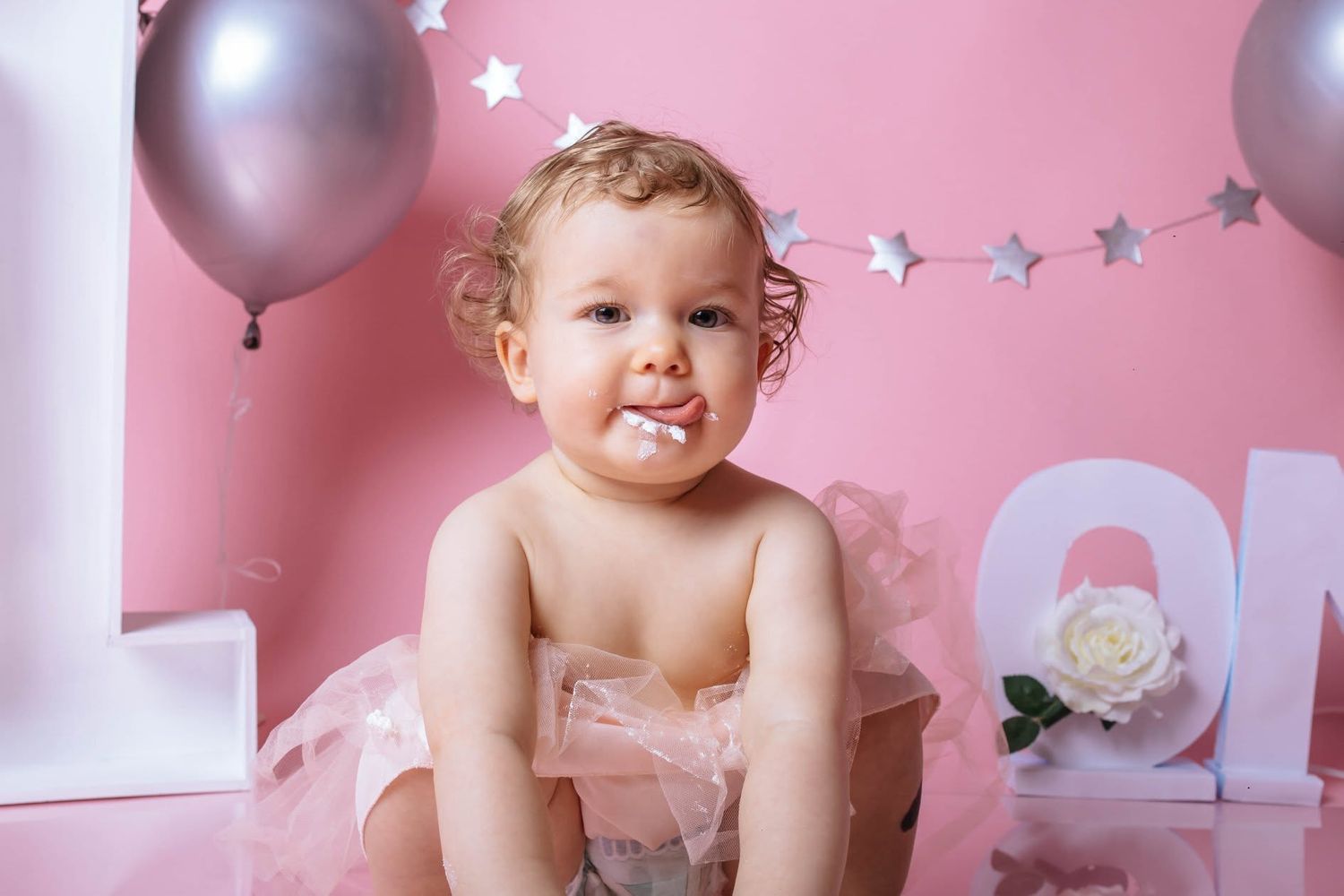 Cute baby girl in pink tutu with cake on her face celebrates first birthday with balloons and letter decorations.