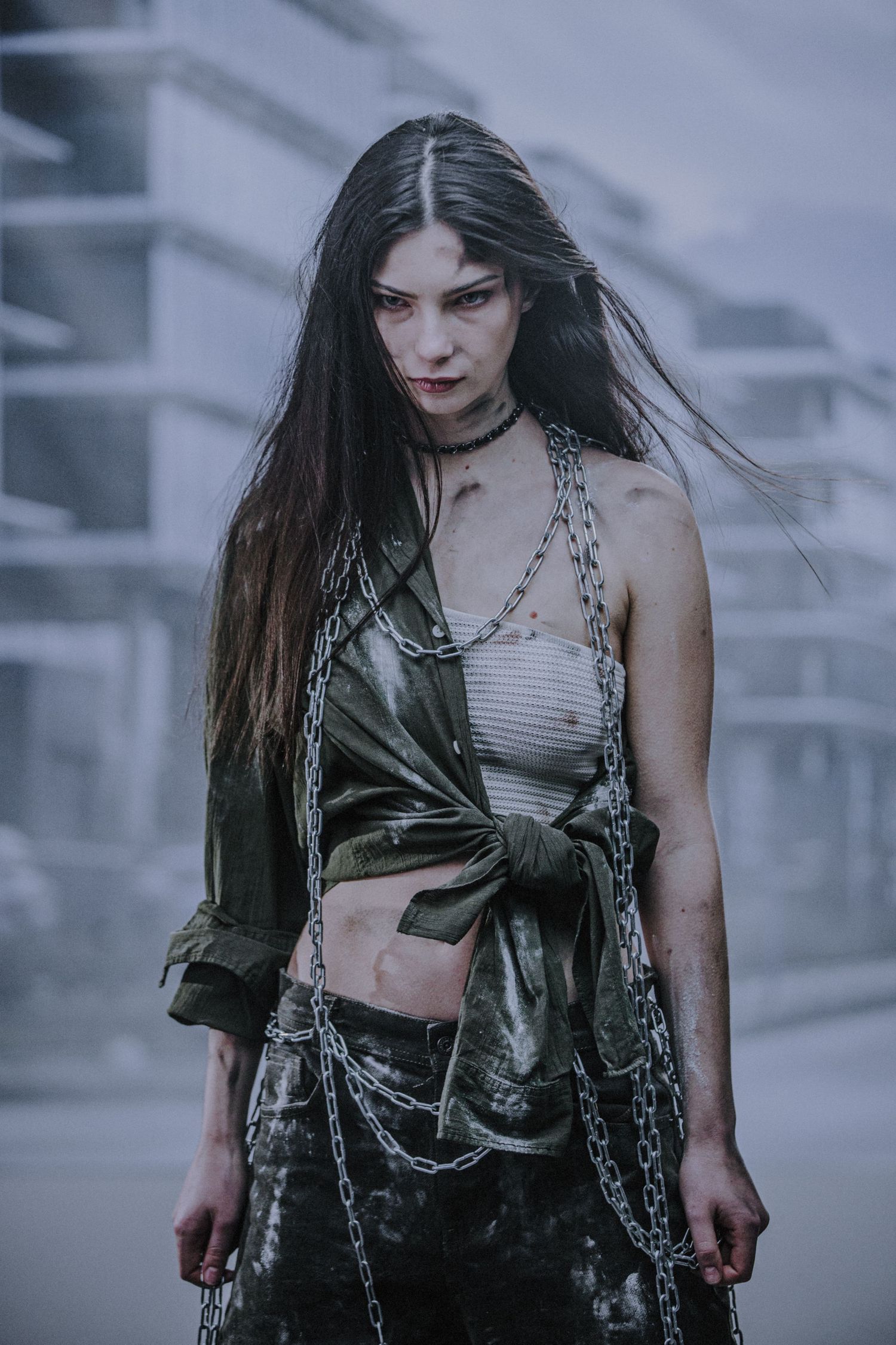 Dark-haired woman in distressed outfit wrapped in metal chains posing in a misty abandoned urban area.