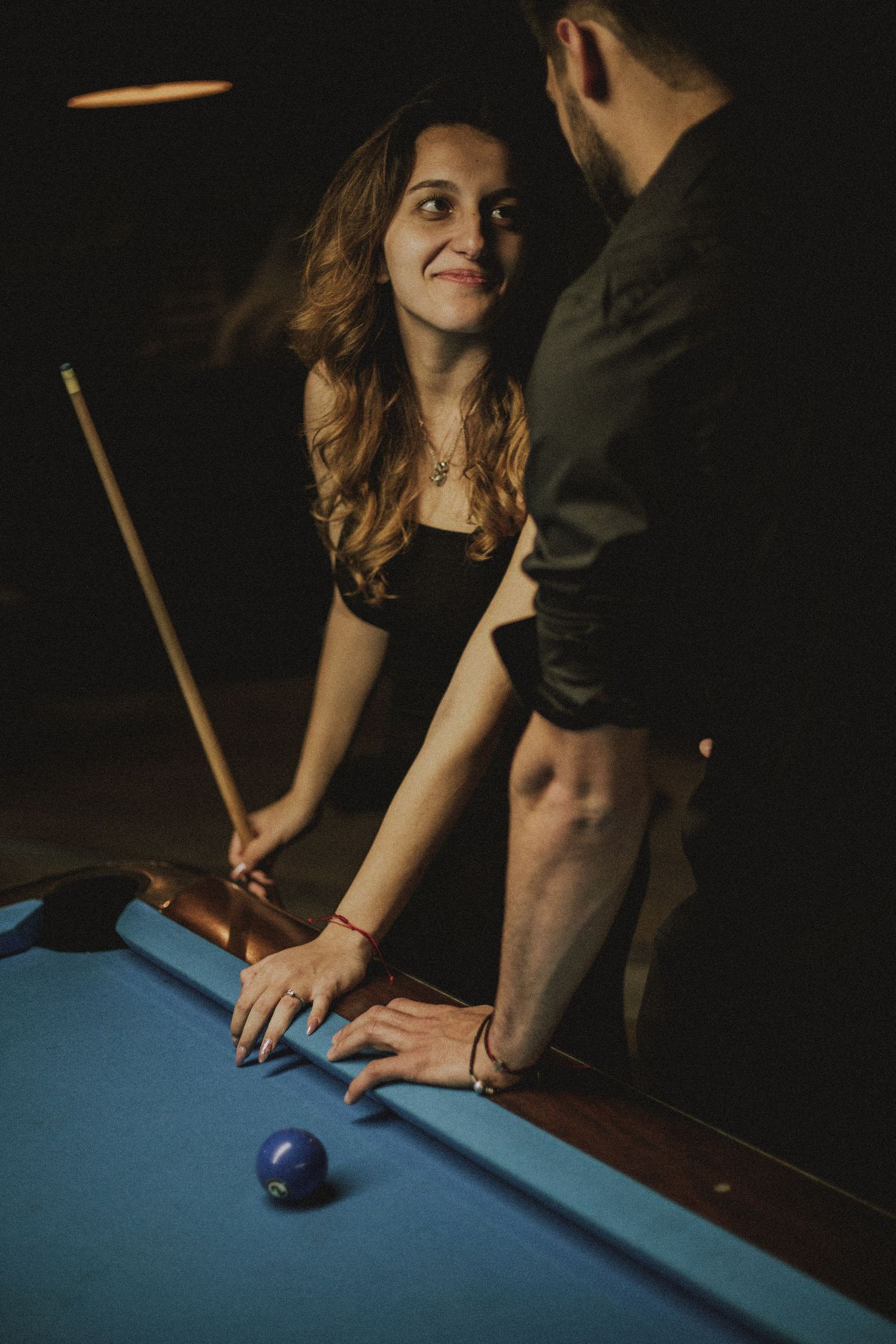 Woman smiling at man while leaning over a pool table holding a billiard cue in a dark bar.