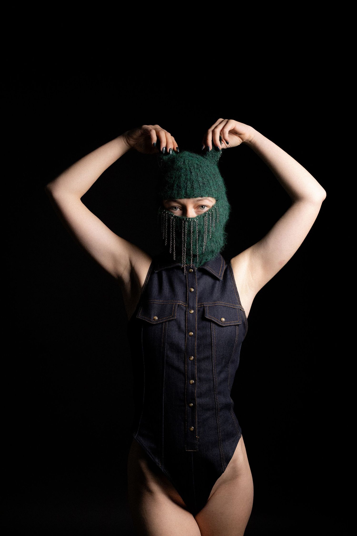 Woman in denim bodysuit wearing green knit balaclava with chains on black background.