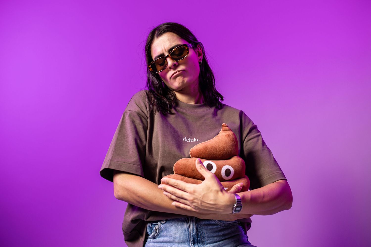 Woman in brown tee and sunglasses hugging a poop emoji plush toy against a purple background.