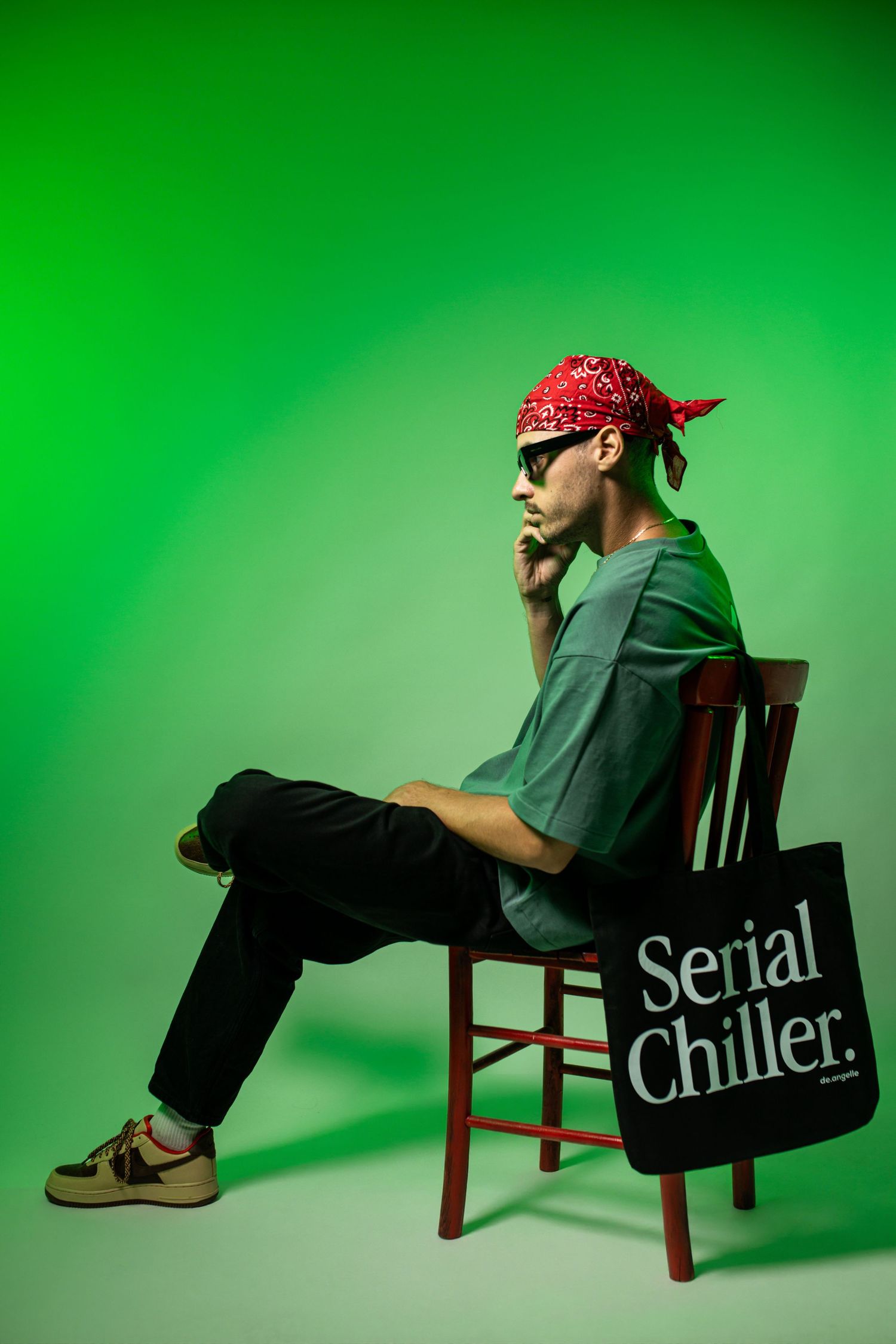 Man in red bandana and sunglasses sitting on chair with Serial Chiller tote bag against green background.
