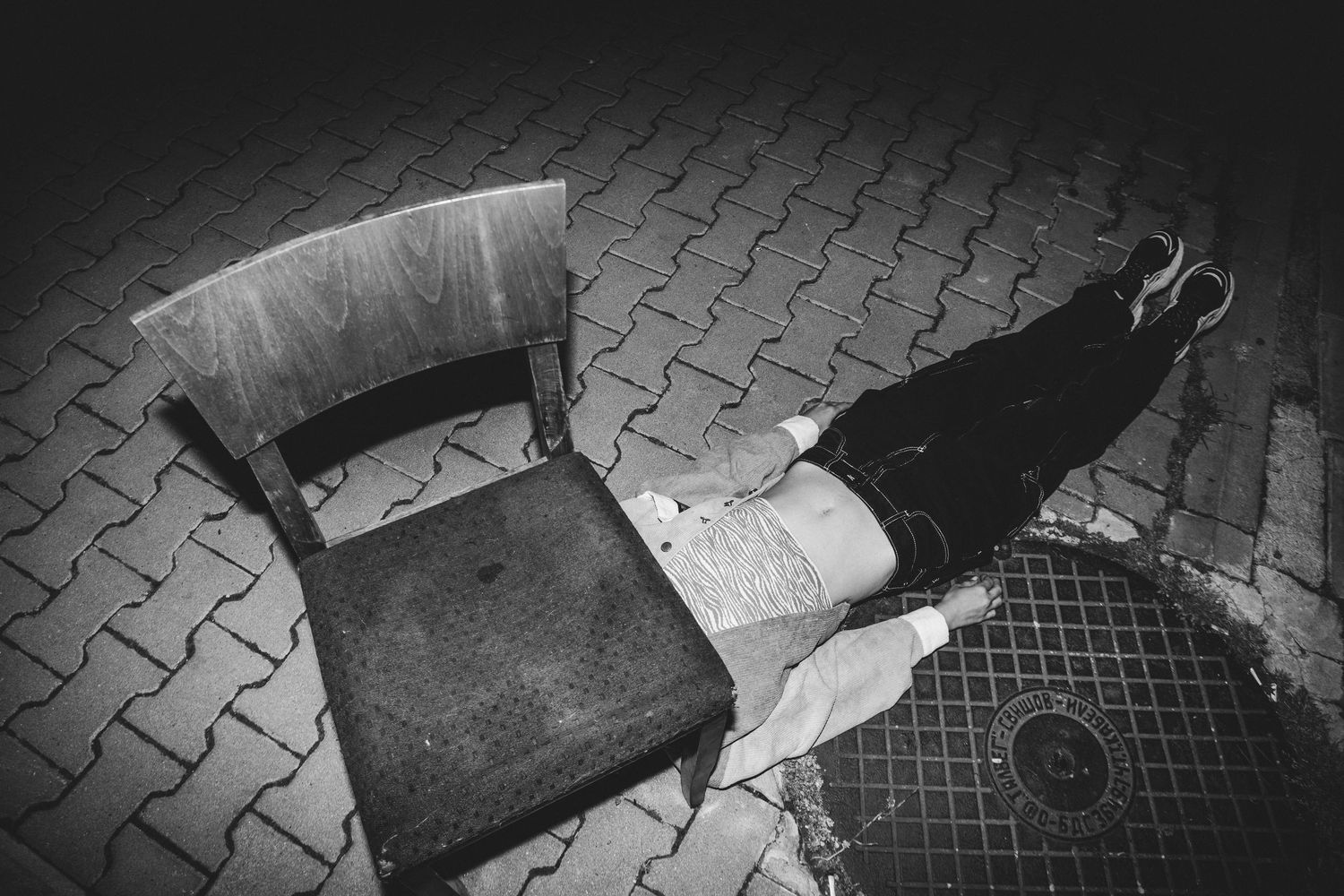 Black and white artistic photo of a wooden chair and persons legs on cobblestone pavement.