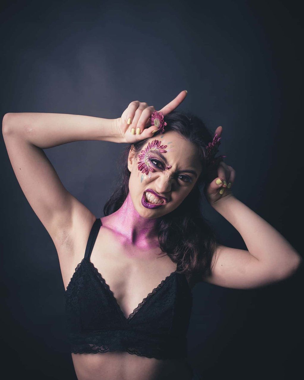 Woman with dramatic pink devil face paint and horns posing in black lace bra on dark background.