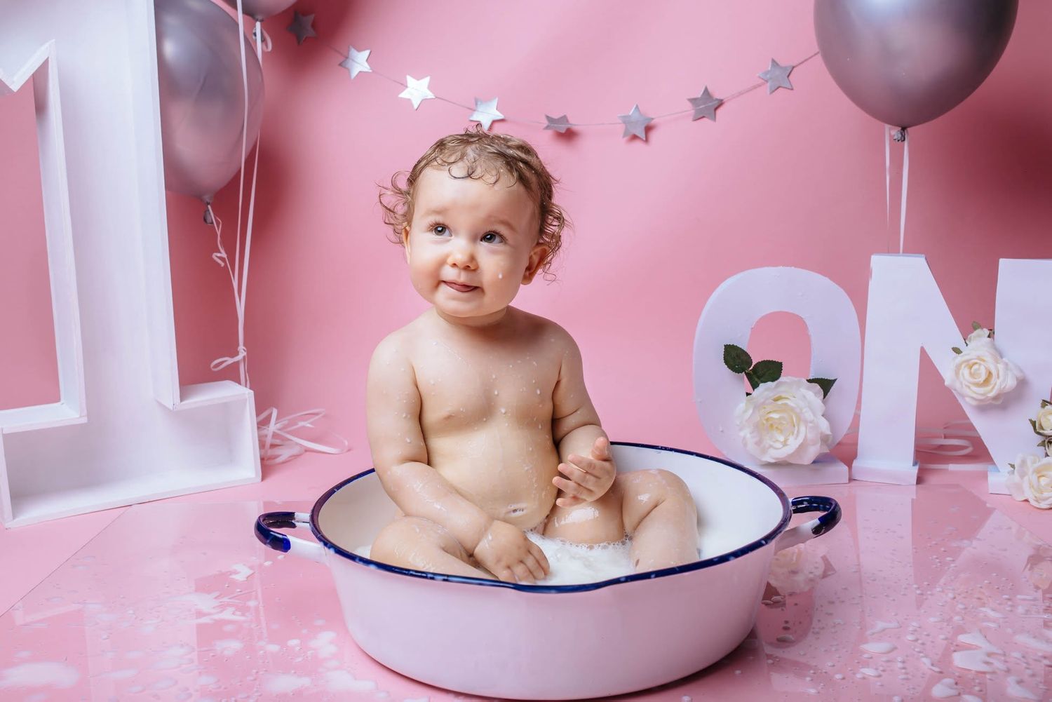 Baby sitting in a small tub during a first birthday cake smash photo shoot with pink decorations.