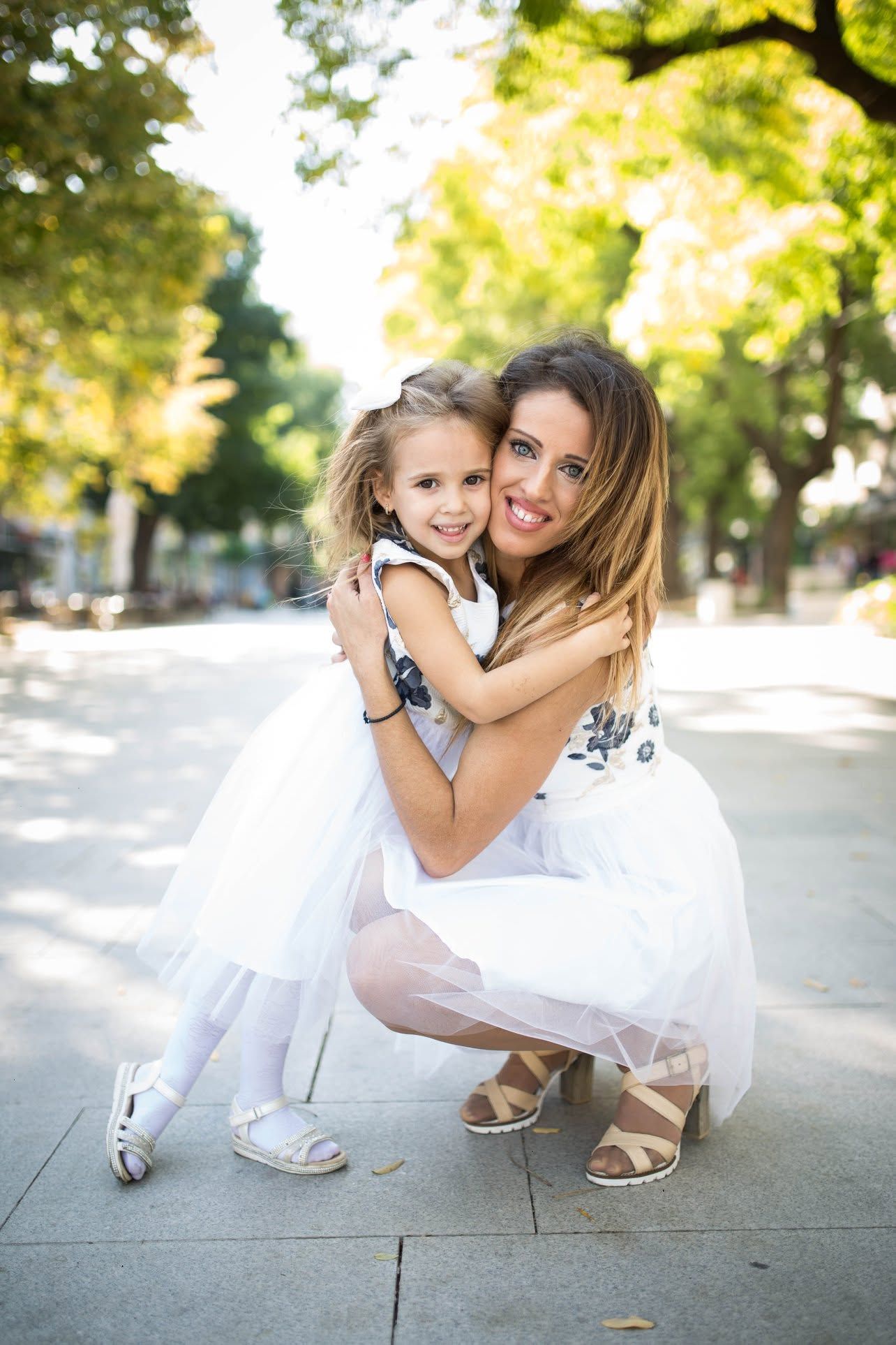 A woman and young girl in matching white dresses embrace and smile outdoors in a sunny park setting.