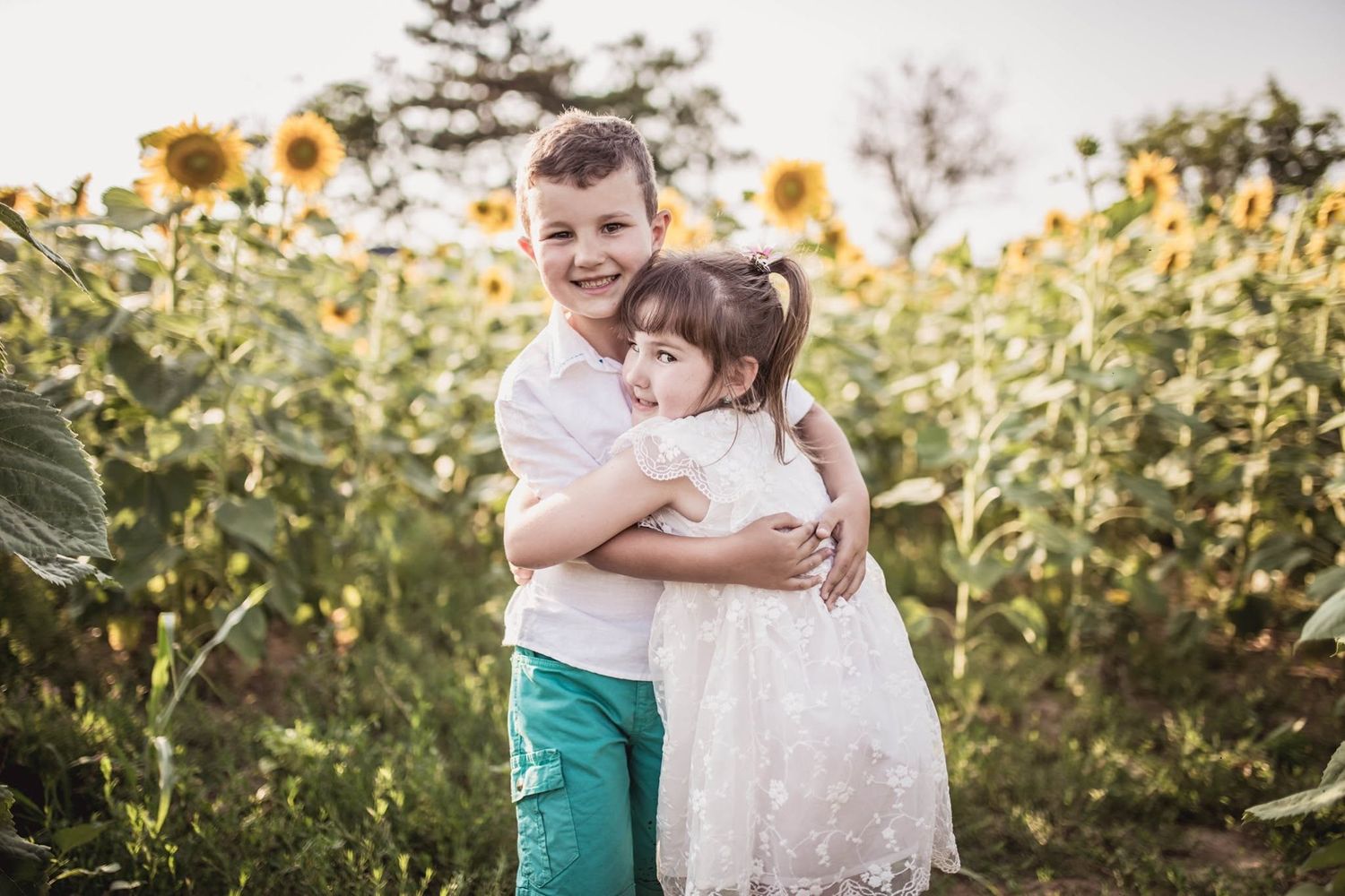 Two children hugging in a sunflower field, boy in teal pants and girl in white dress, smiling outdoors.