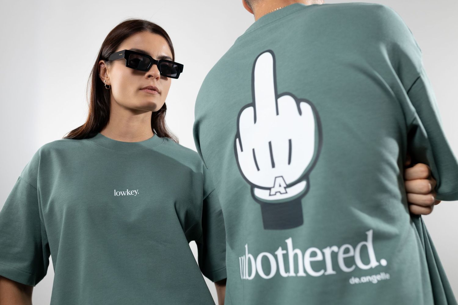 Two models wearing sage green oversized t-shirts with 'unbothered' graphic print and middle finger logo design.