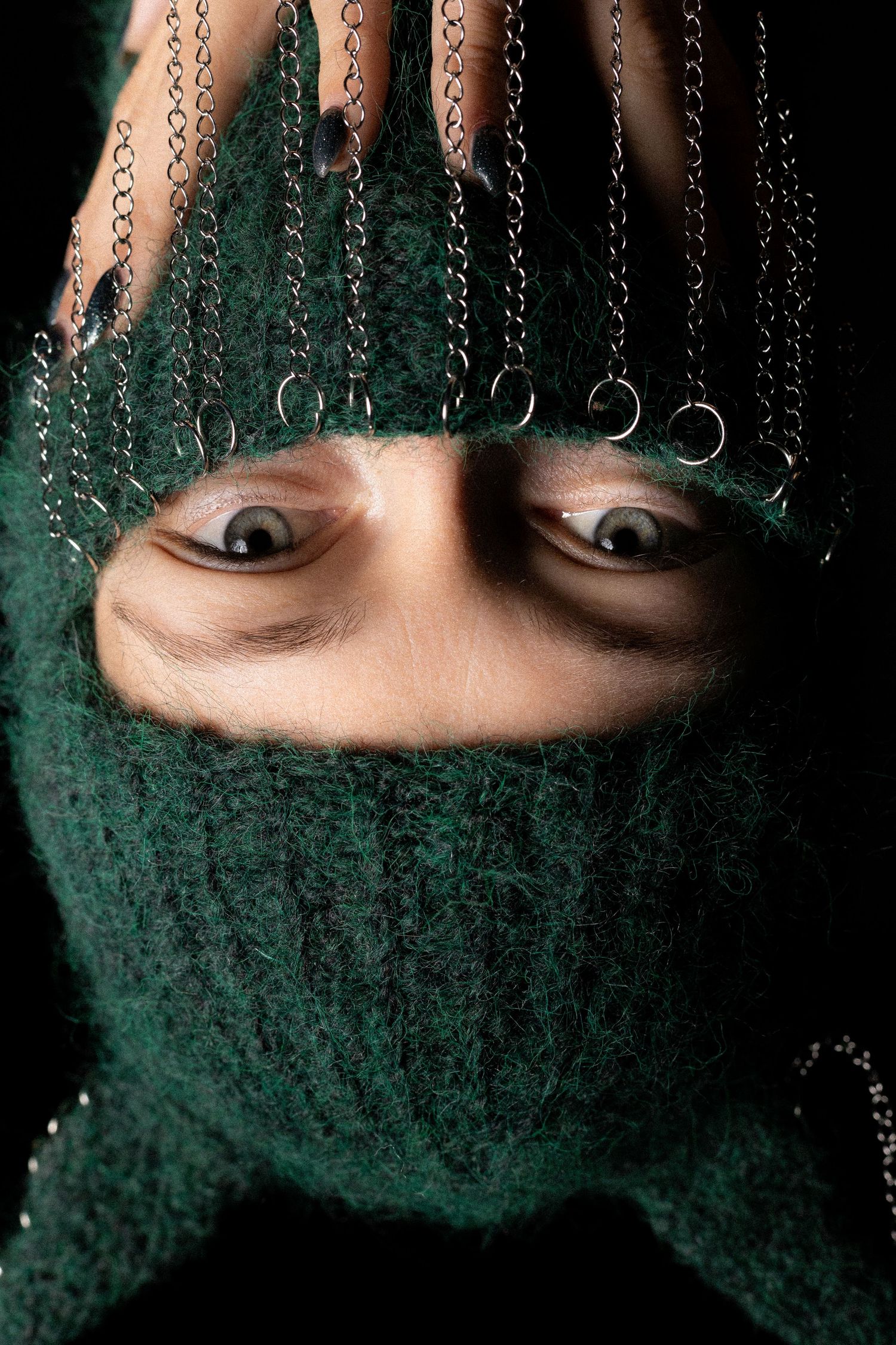 Close-up of intense eyes through green knit balaclava with decorative metal chains.