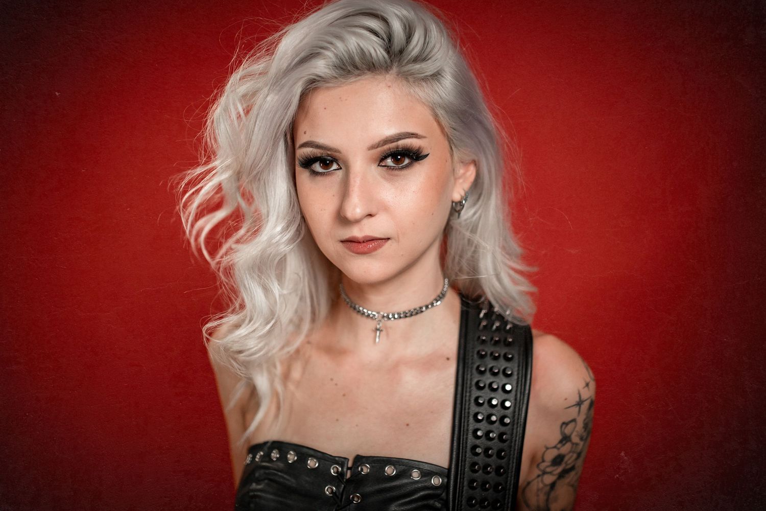 Young woman with silver wavy hair, tattoos, and black studded corset posing against red background.