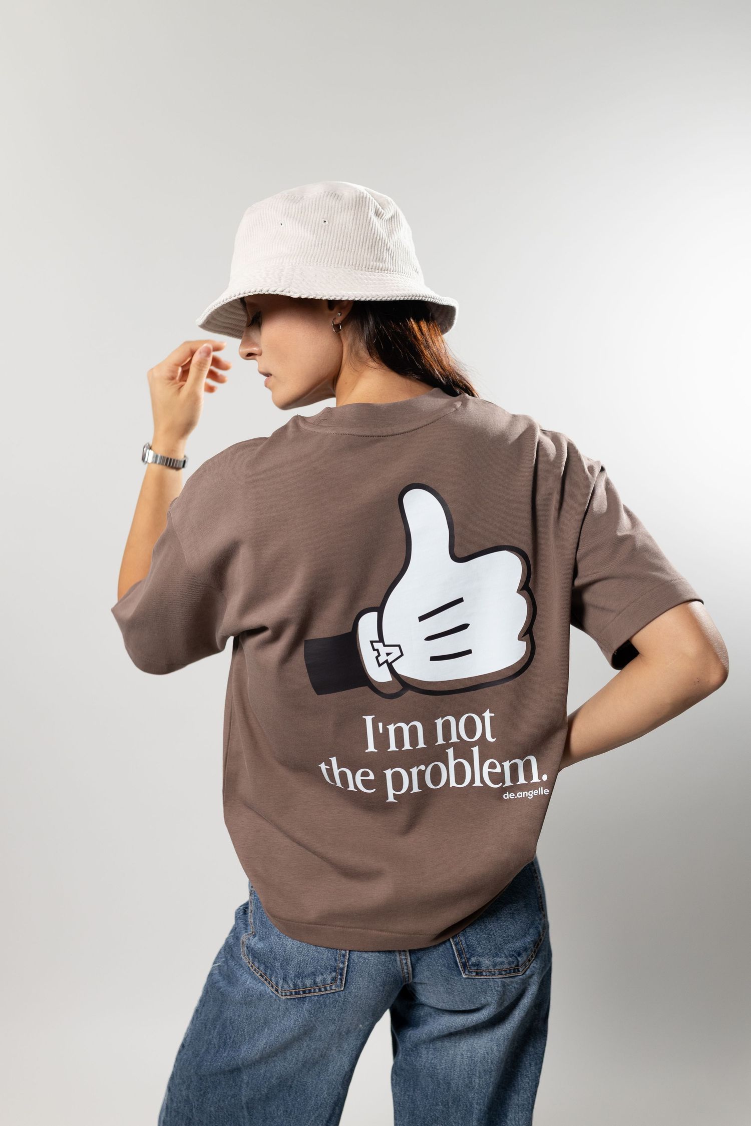 Woman wearing brown oversized graphic tee with thumbs up print and 'I'm not the problem' text, paired with jeans and bucket hat.