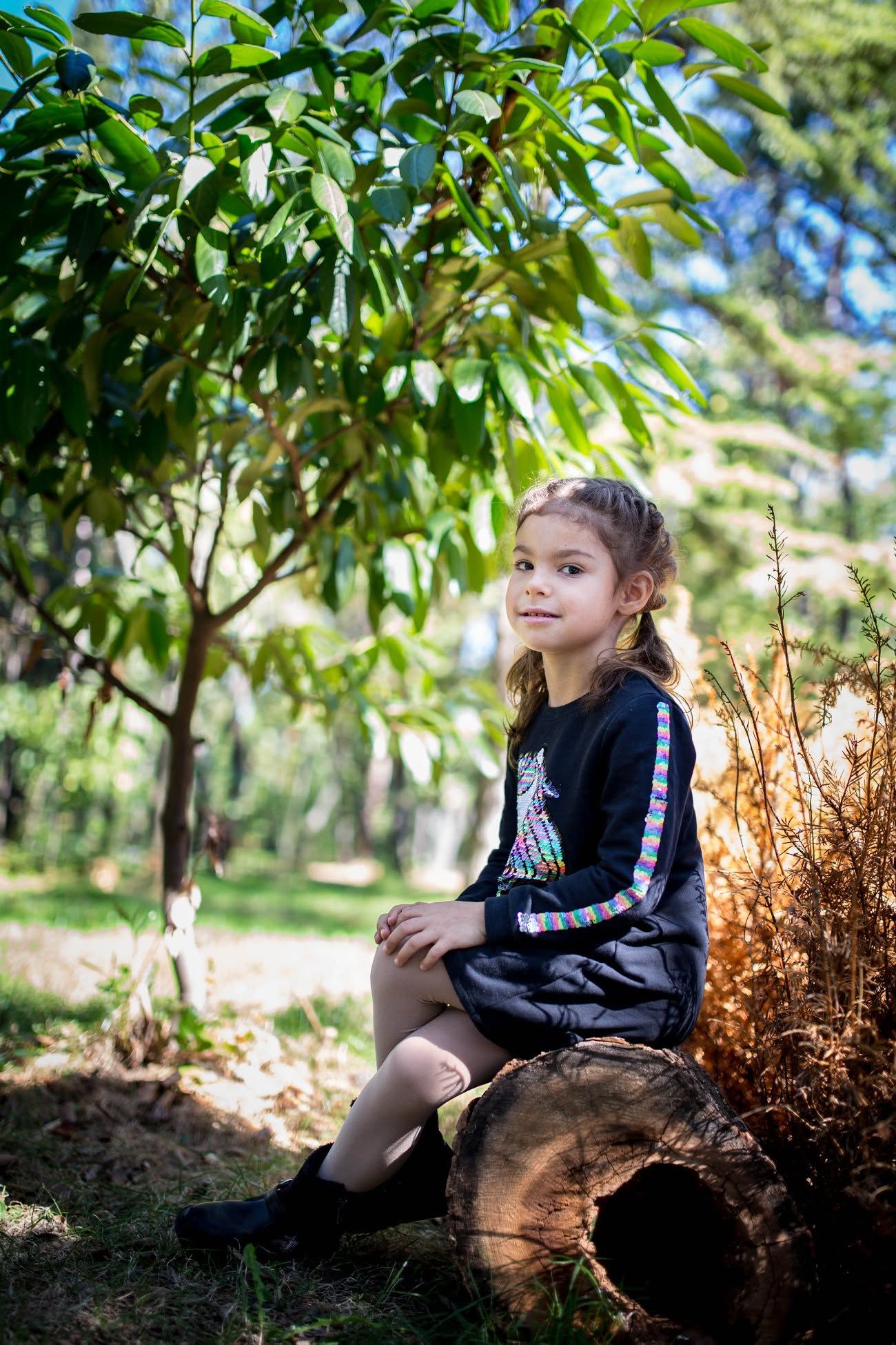 Young girl in black outfit sitting on a log in a lush green park, smiling softly at the camera.