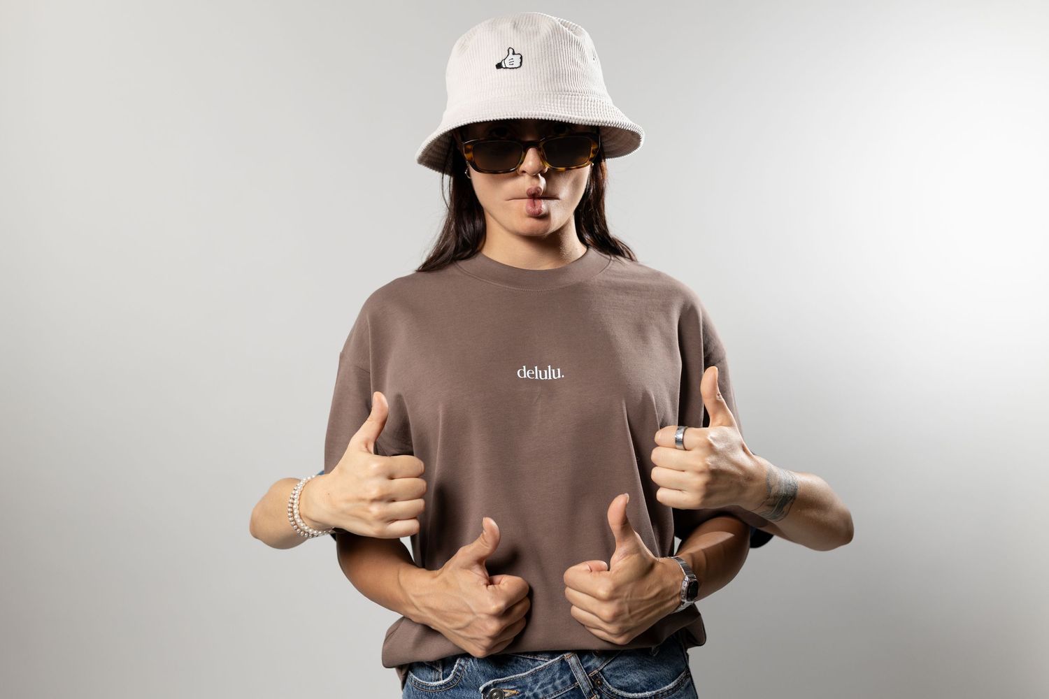 Woman in brown oversized tee, white bucket hat, and sunglasses giving thumbs up with multiple arms effect.