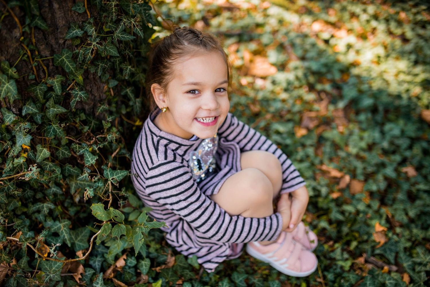 A smiling young girl in a striped sweater sits outdoors among green ivy leaves and autumn foliage.