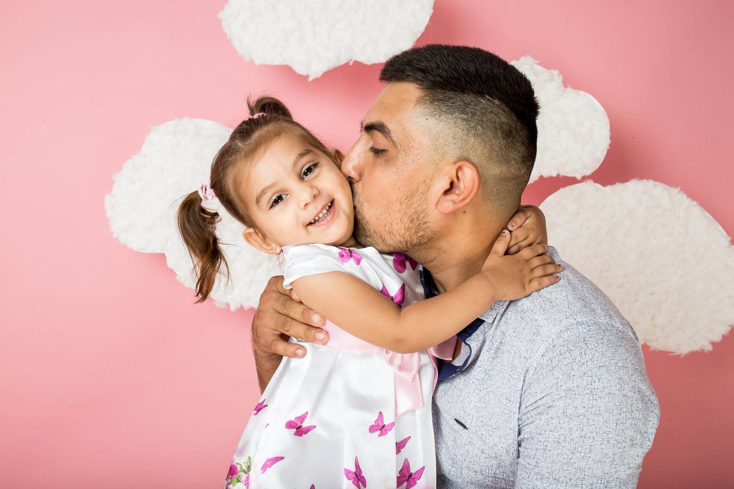 Father kissing his little daughter on the cheek against a pink background.
