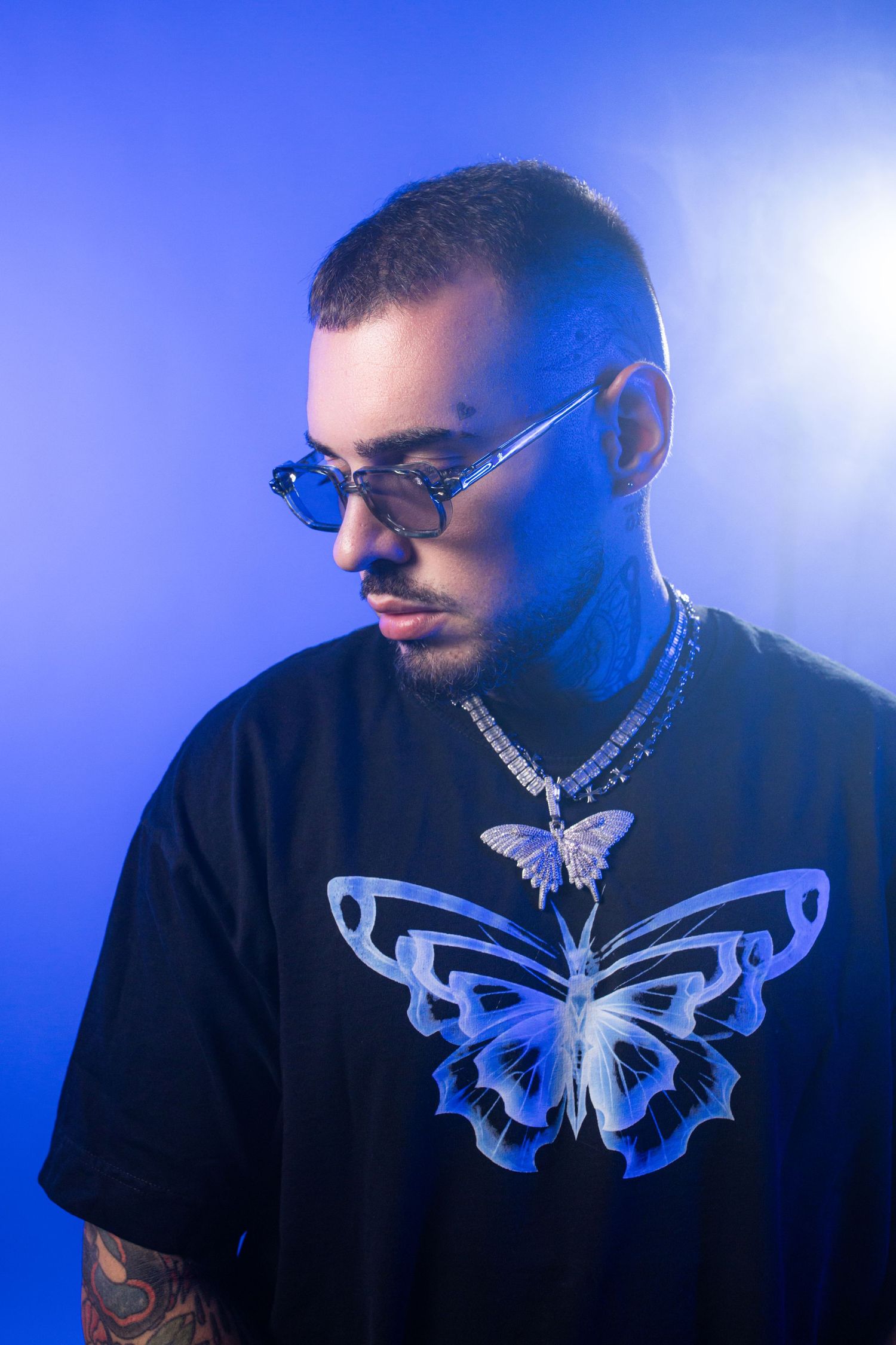 Man in black butterfly t-shirt, blue sunglasses, diamond necklace, and tattoos poses against a blue background.