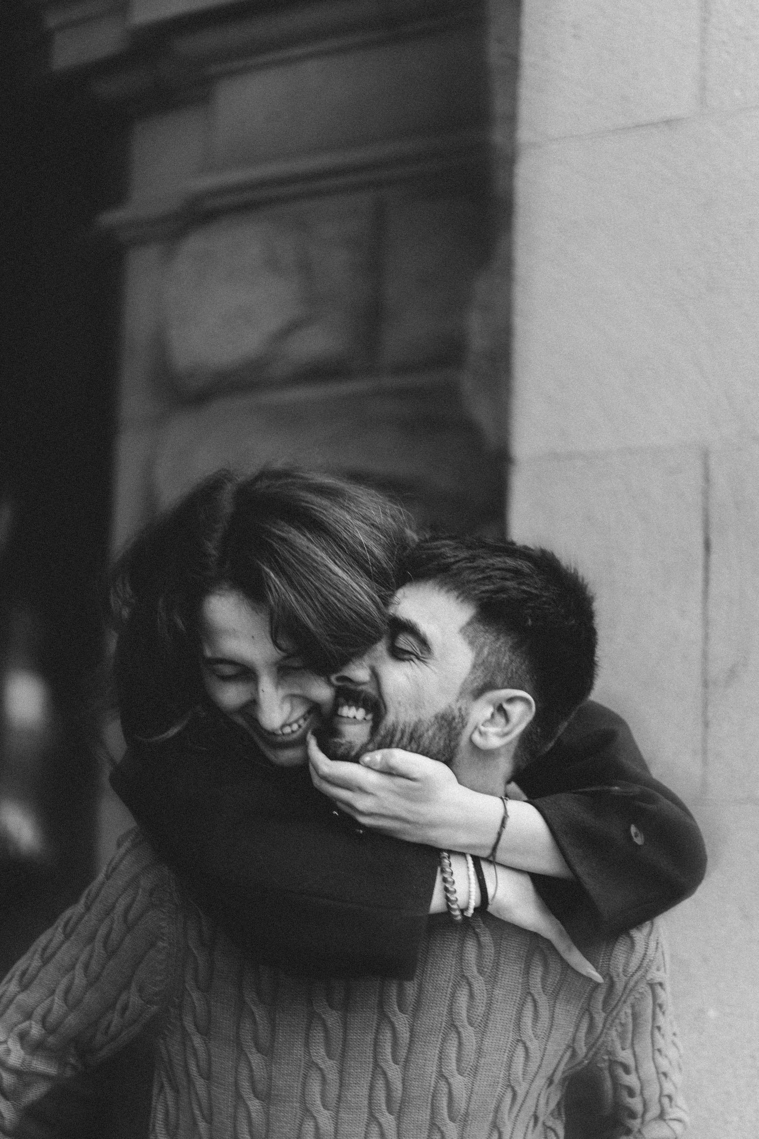 Black and white photo of a happy couple hugging and laughing outdoors against a stone wall.