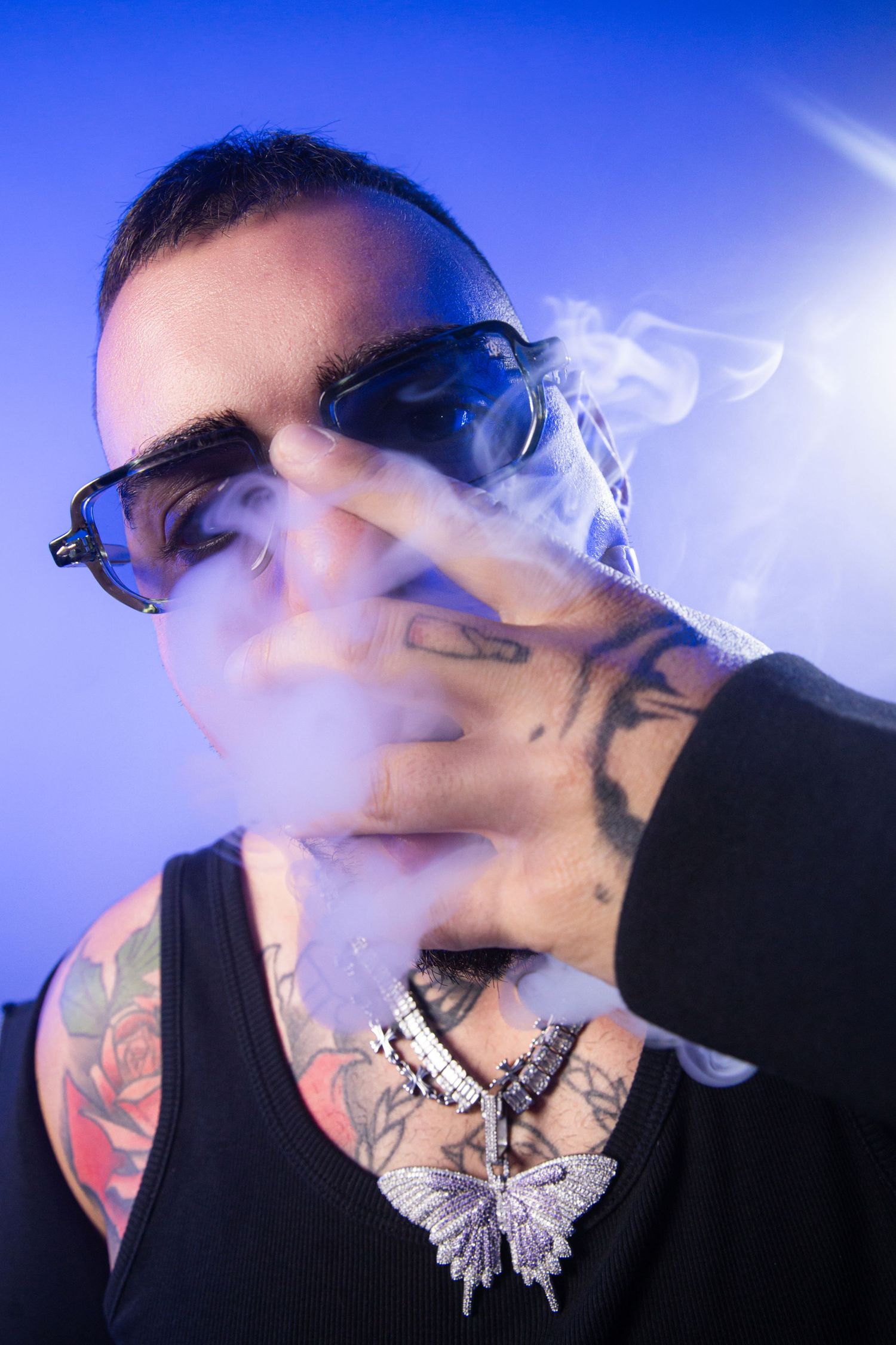 Tattooed man with sunglasses exhaling smoke, wearing butterfly necklace and black tank top against blue background.