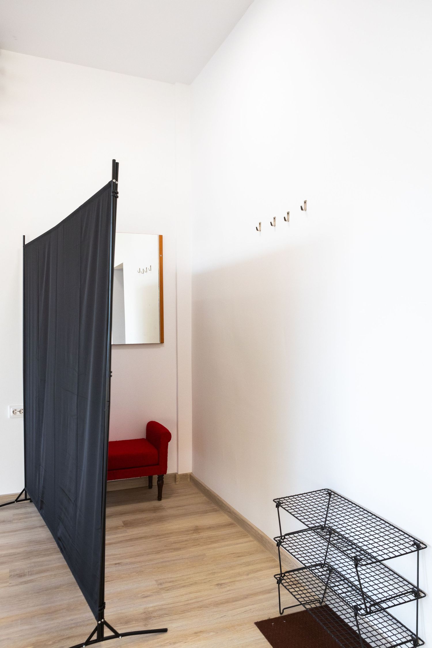 Minimalist hallway with dark fabric room divider, red accent chair, wire shoe rack, and coat hooks.