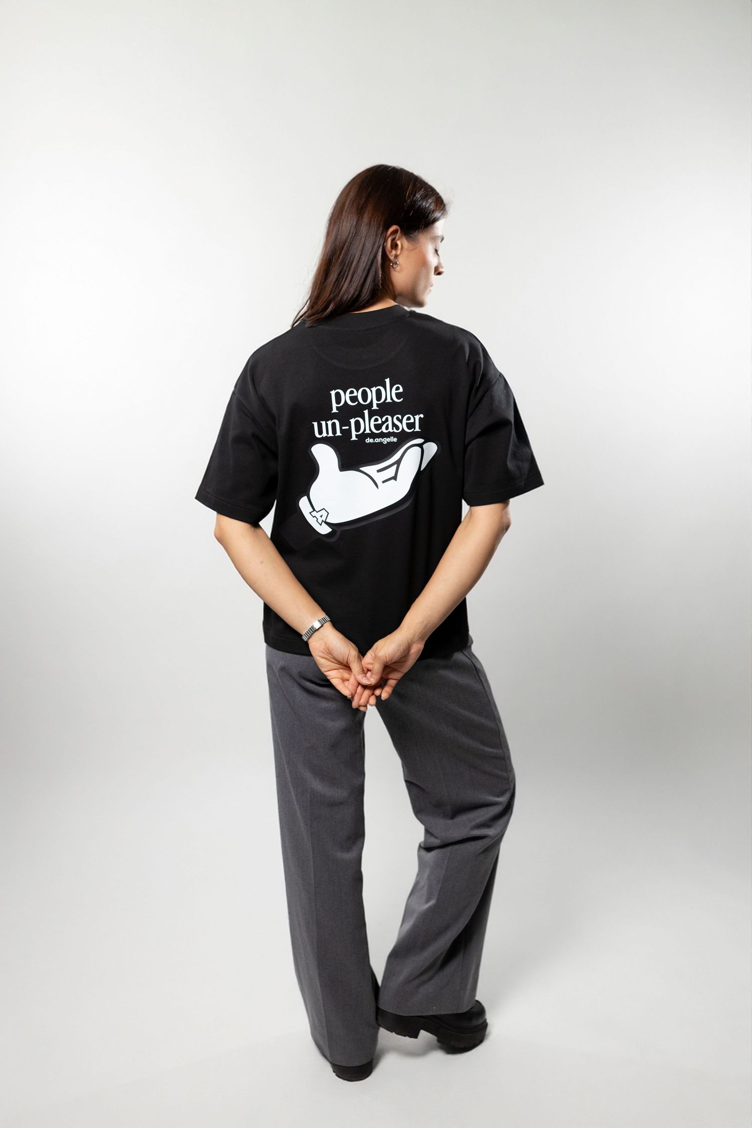 Woman wearing black oversized 'People Un-Pleaser' graphic t-shirt with grey wide-leg pants, shown from behind.