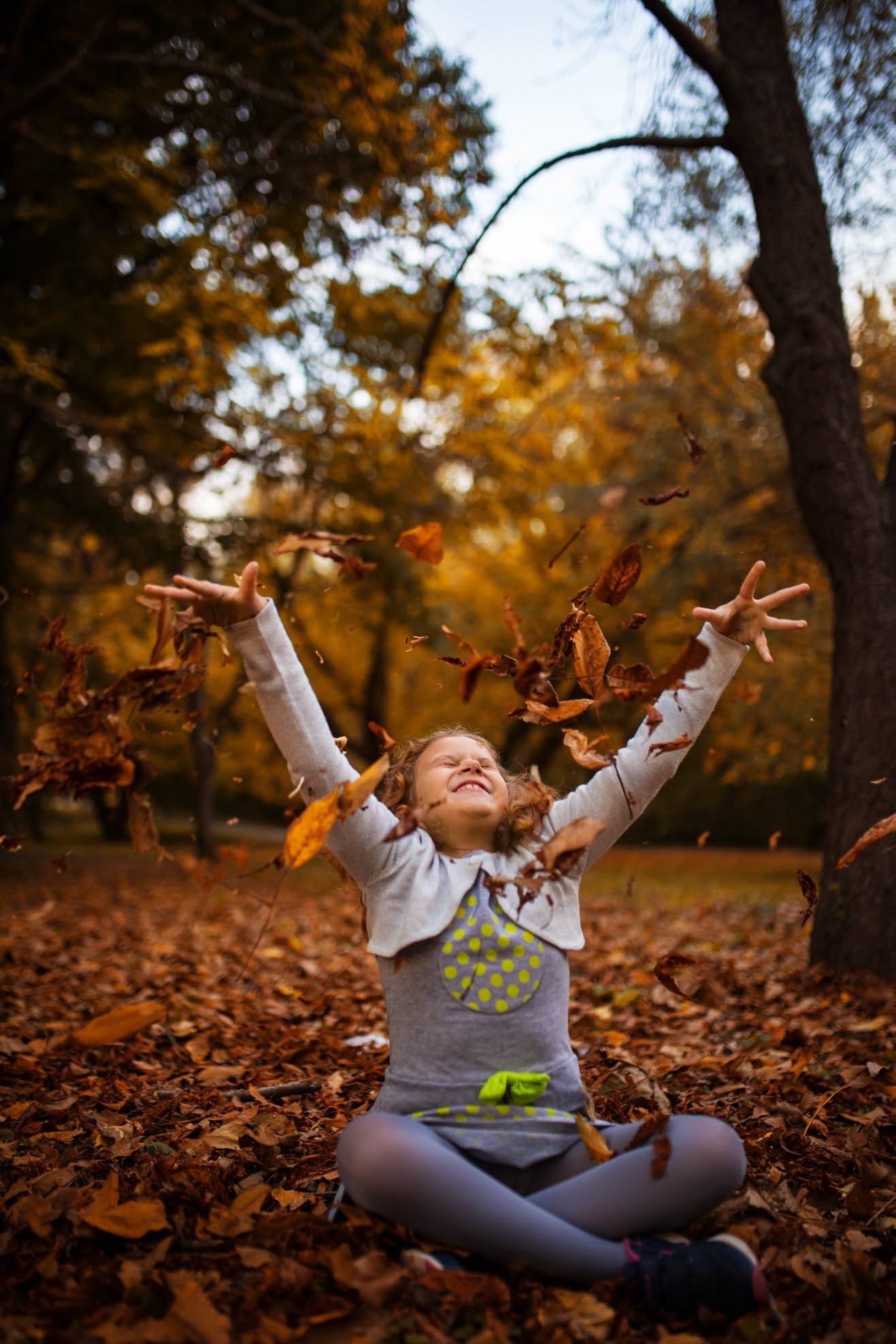 A joyful girl sits cross-legged in an autumn park, tossing golden fallen leaves into the air with arms raised high.