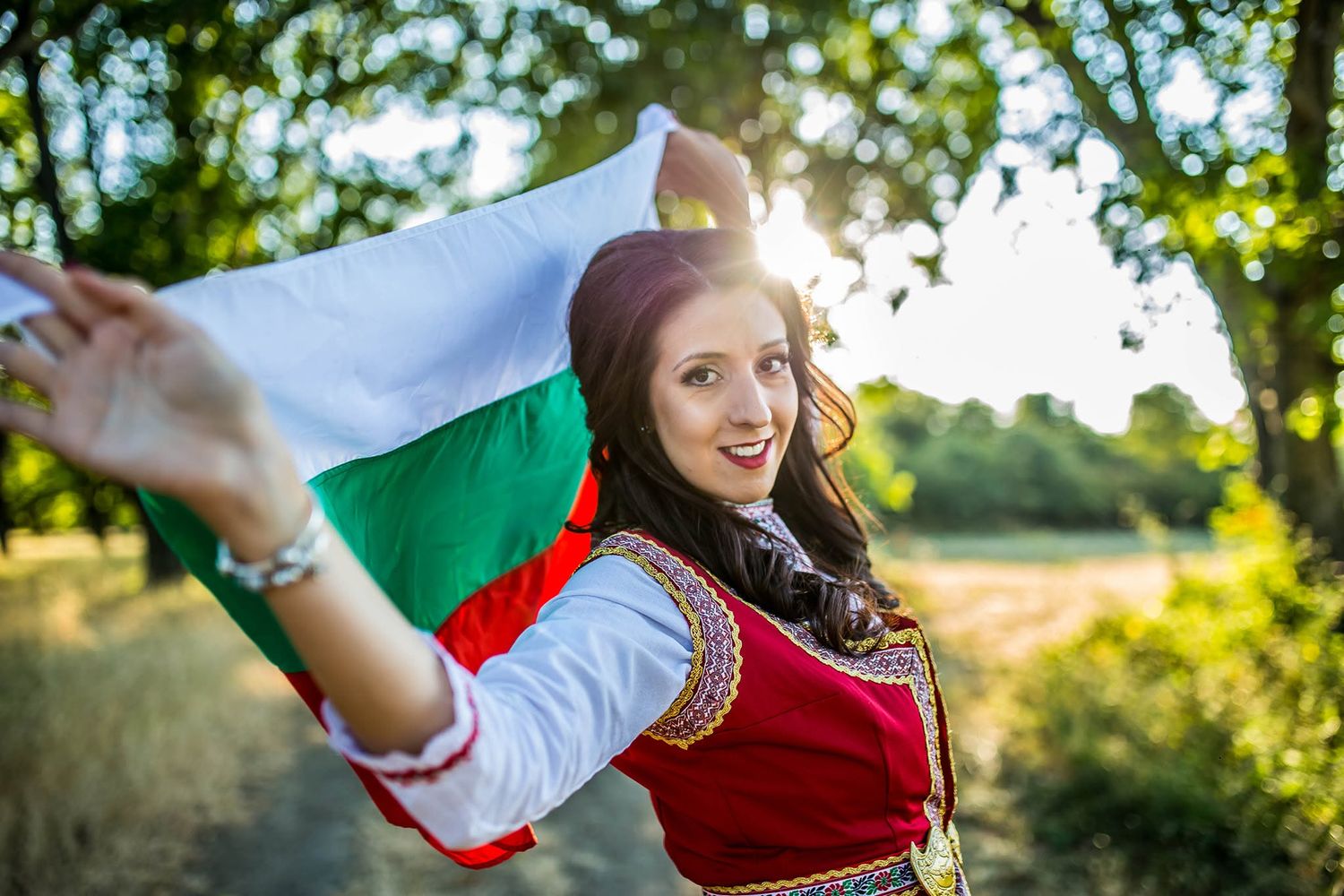 Young woman in traditional Bulgarian folk costume smiling while holding a Bulgarian flag outdoors in golden sunlight.