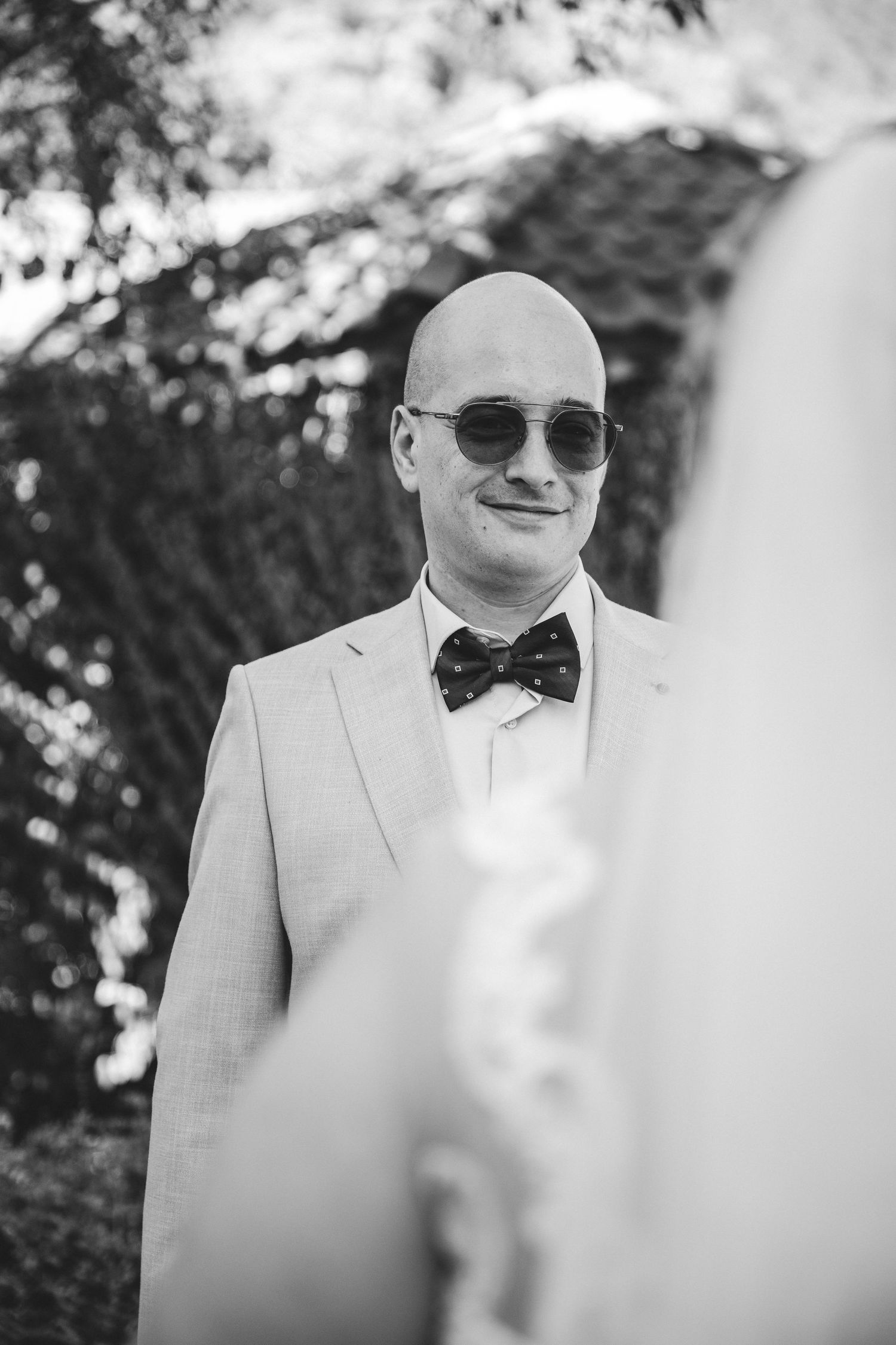 Bald groom in light suit and polka dot bow tie wearing sunglasses smiles at bride during outdoor wedding ceremony.