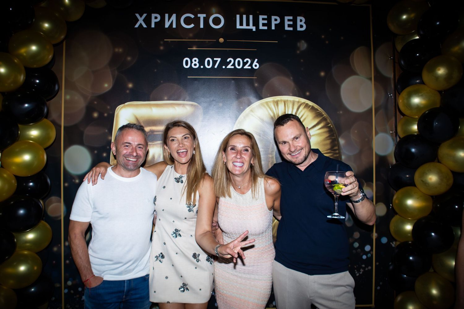 Four people smiling at a birthday celebration photo booth with gold and black balloons and a banner dated 08.07.2026.