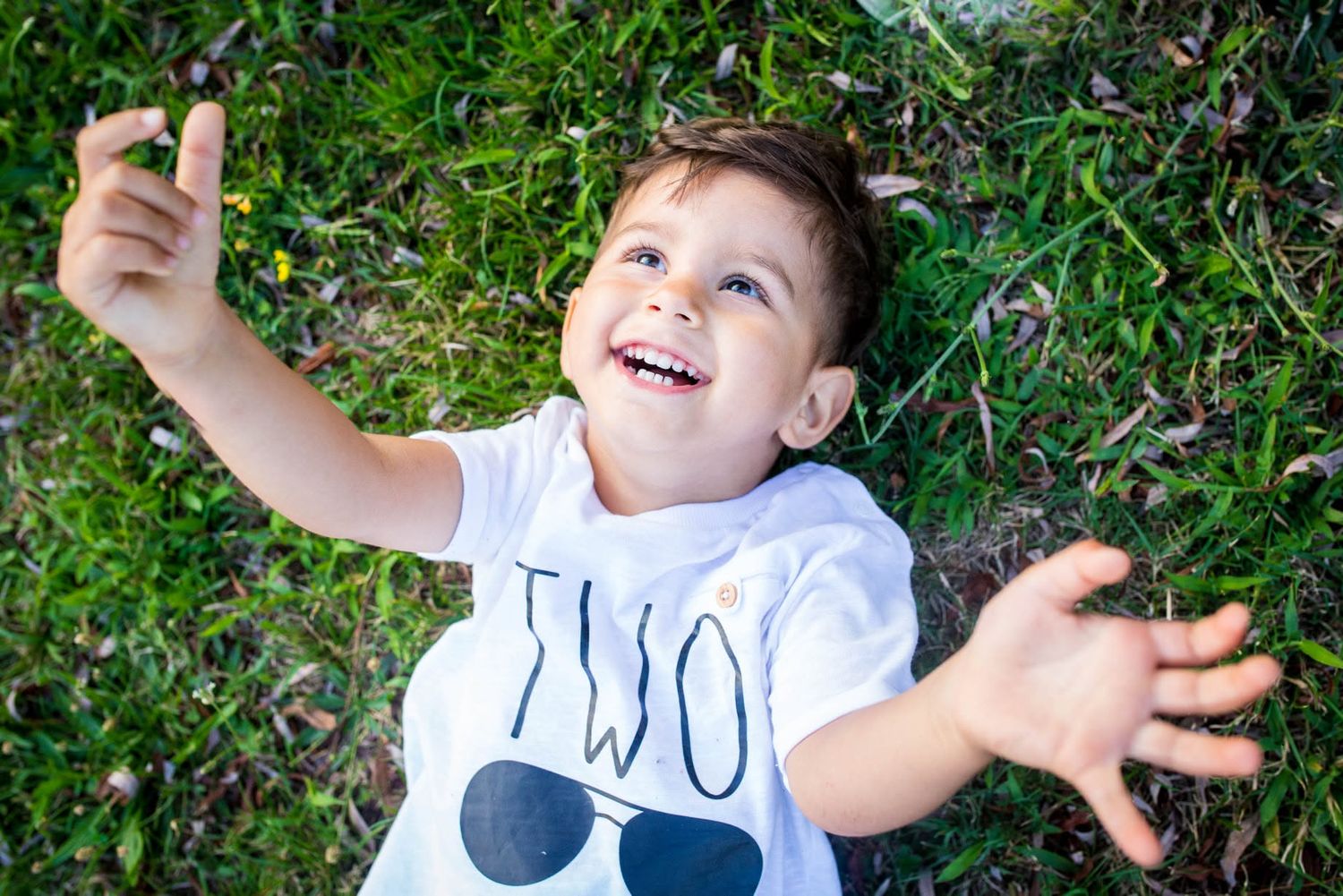 Happy toddler in a white TWO shirt lying on green grass, laughing and reaching arms up in joy.