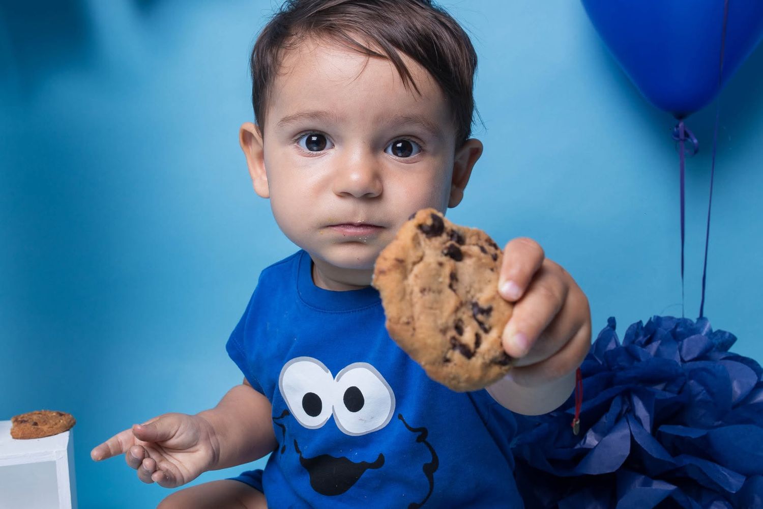 Baby in Cookie Monster shirt holds chocolate chip cookie at first birthday party with blue balloon backdrop.