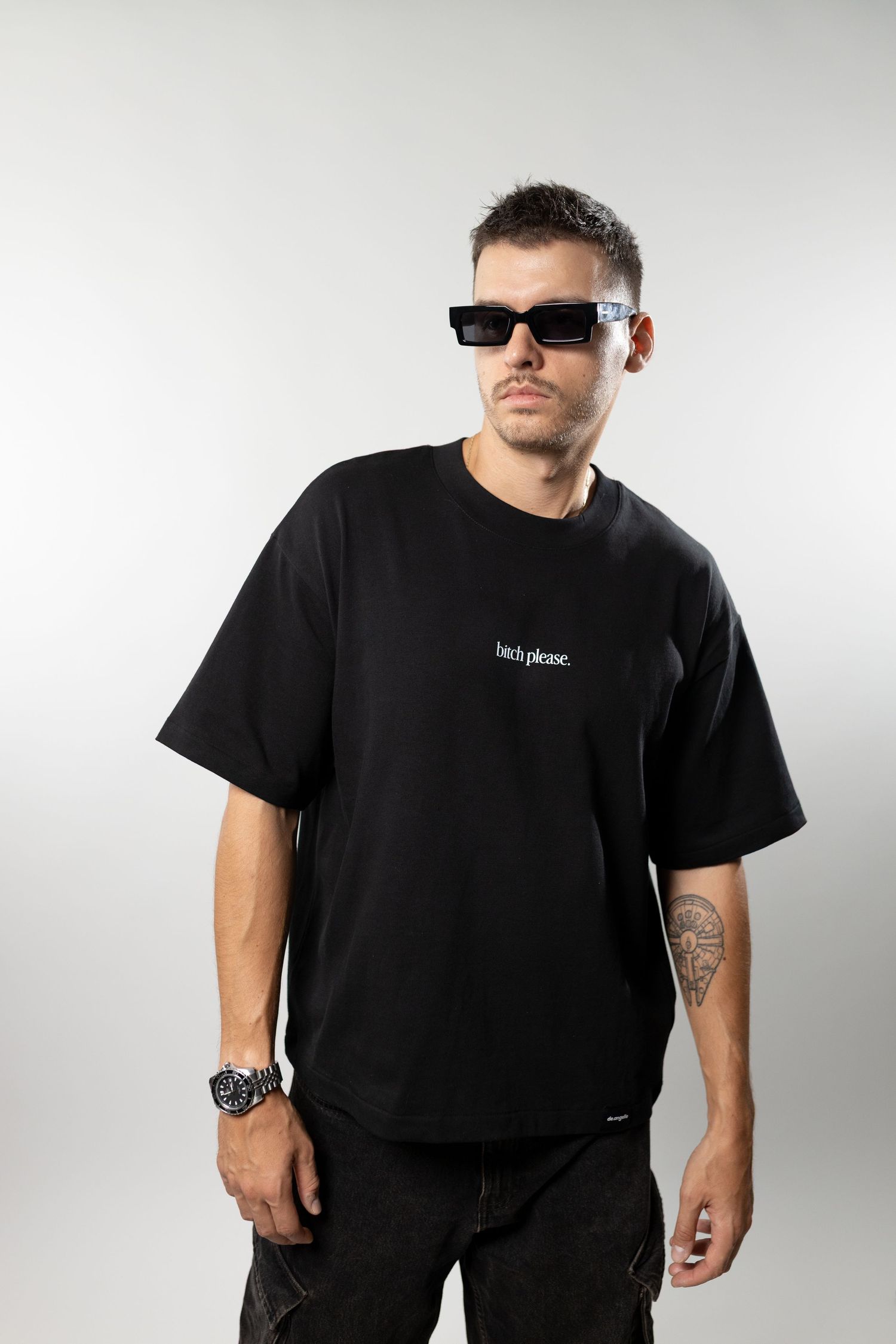 Man wearing black oversized graphic t-shirt with sunglasses and watch, posing against gray background.