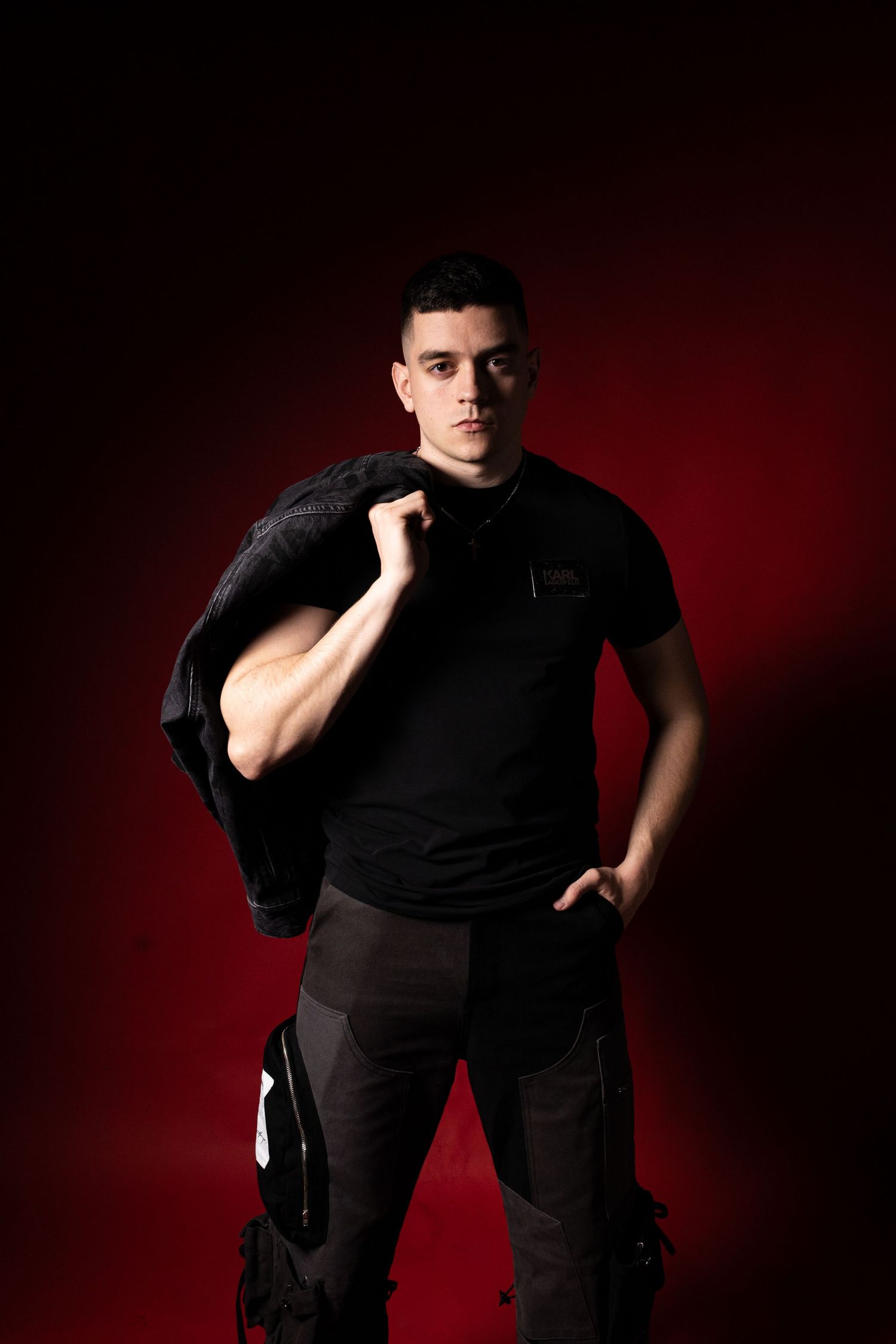 Confident young man in black outfit holding jacket over shoulder on dark red background.