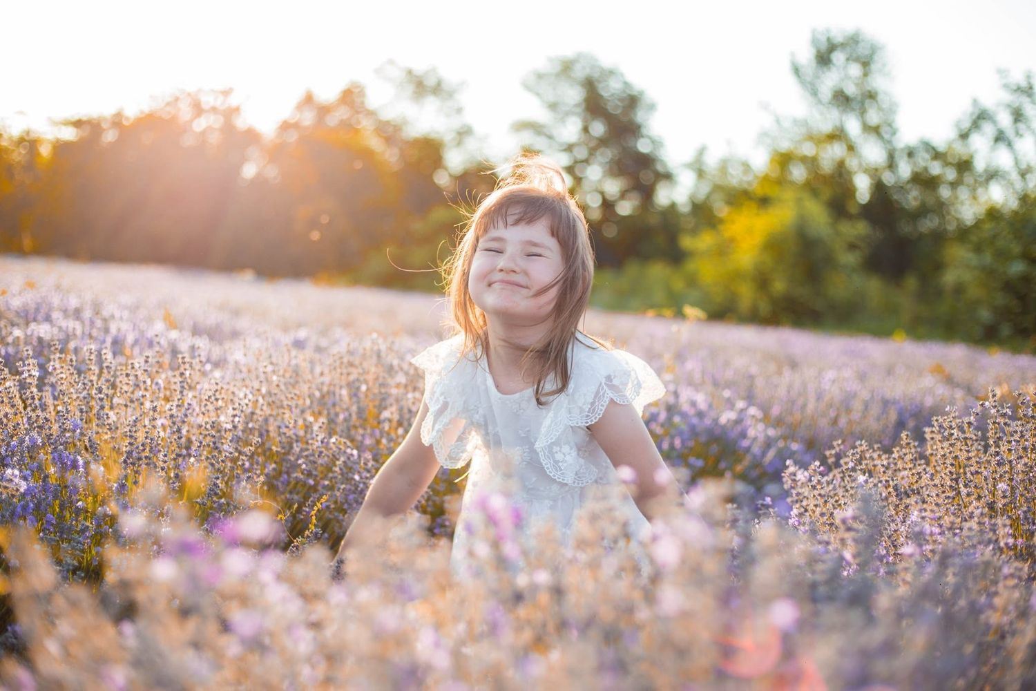 A joyful young girl in a white dress smiles while standing in a blooming lavender field during golden hour sunset.