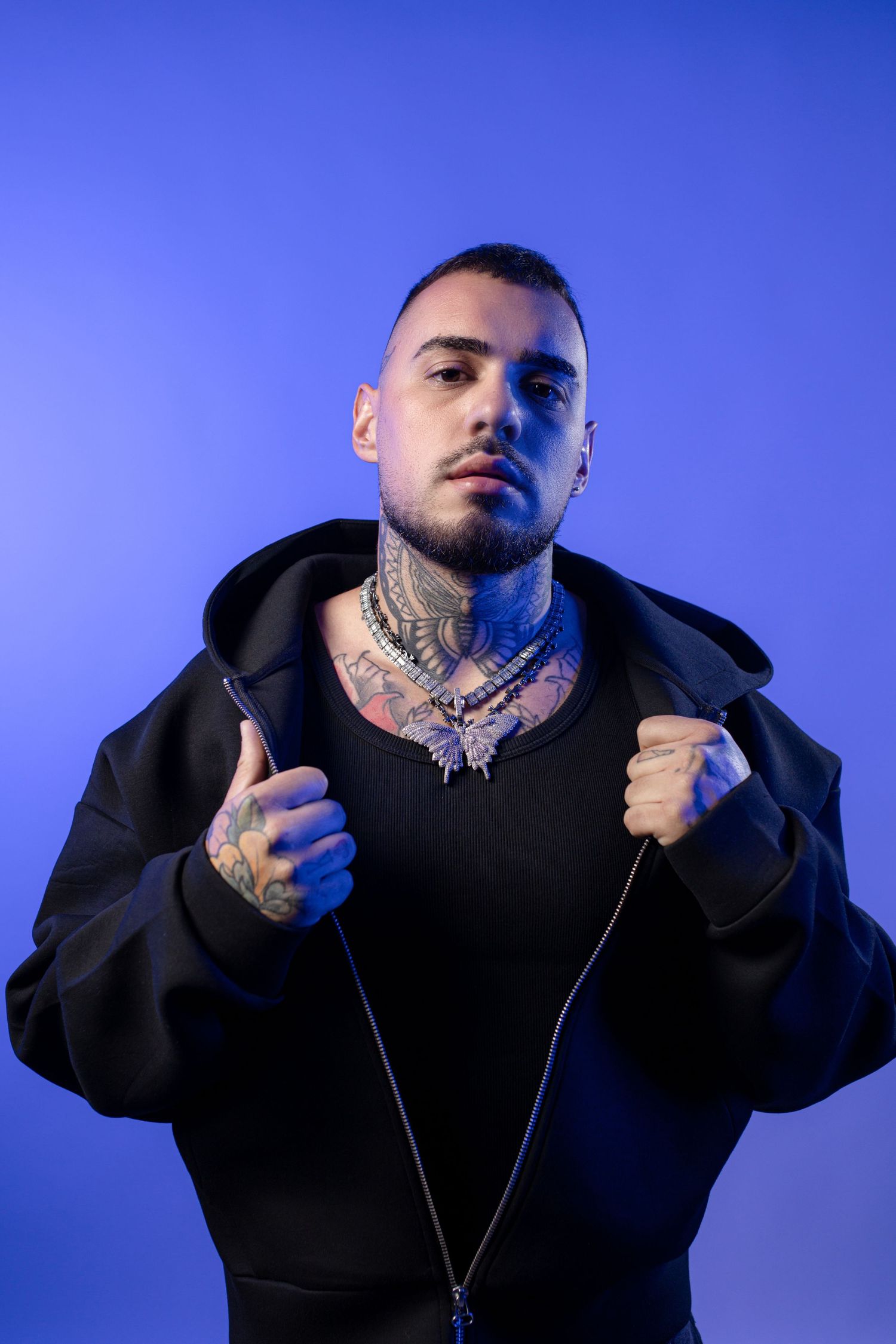 Tattooed man in black hoodie with silver chain necklaces posing against blue background.