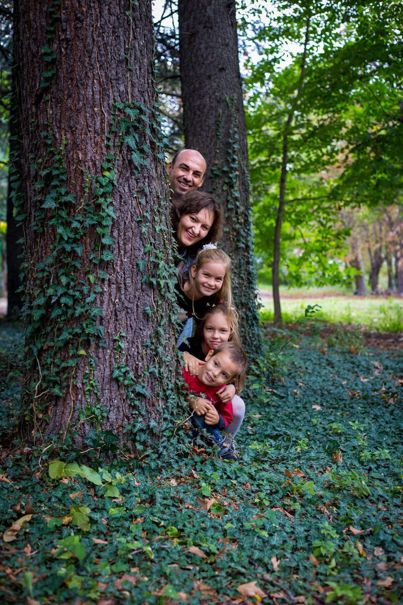 Happy family of four peeking from behind a large tree covered in ivy in a lush green park.