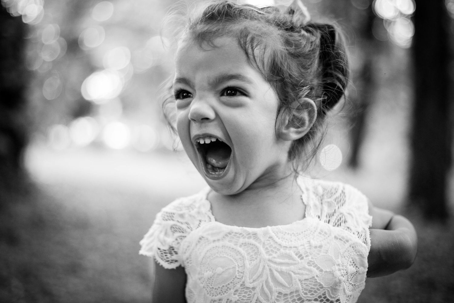 Joyful toddler girl in white lace dress laughing with mouth wide open in black and white photo.