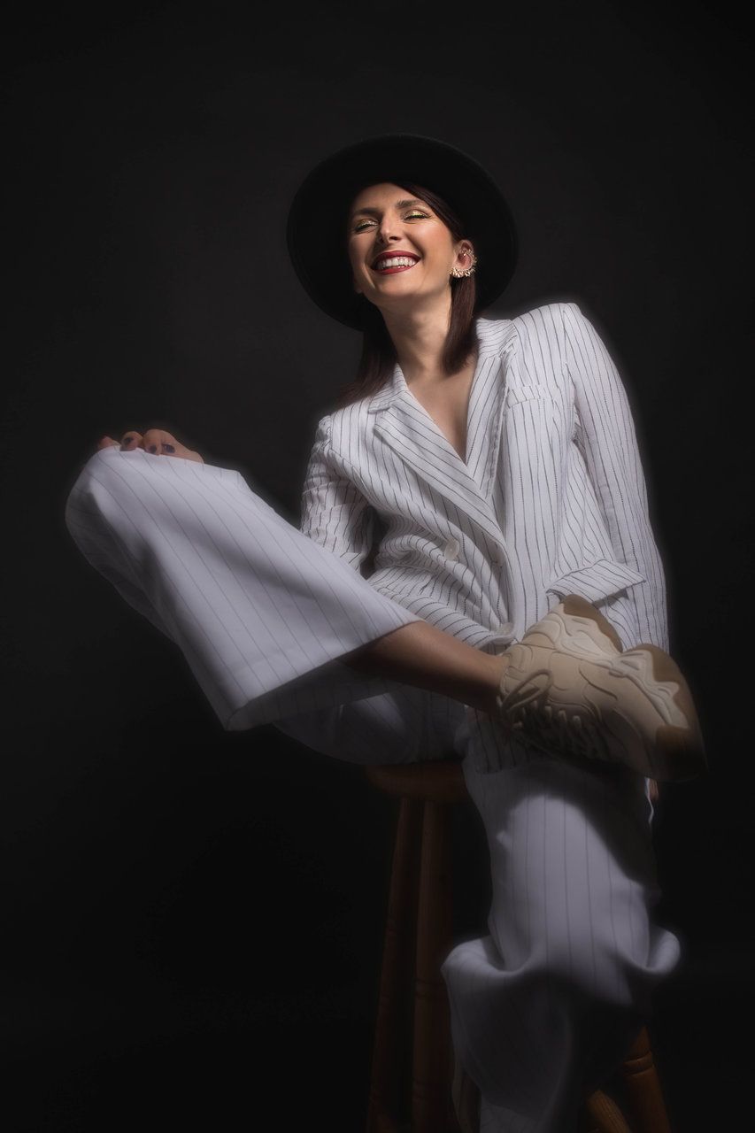 Smiling woman in white pinstripe suit and black wide-brim hat sitting on wooden stool against dark background.