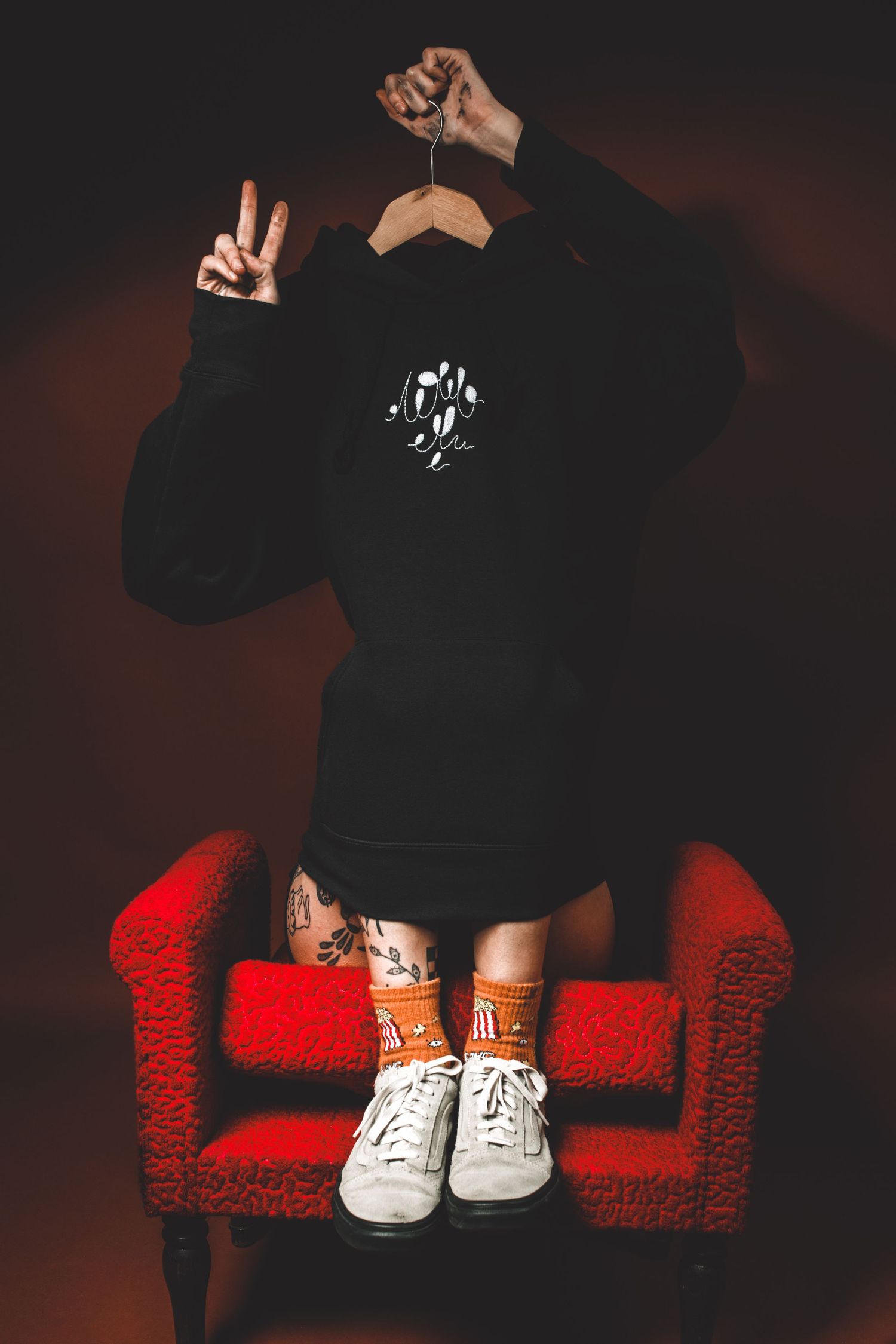 Person sitting on red chair hides face behind black hoodie on hanger, showing peace sign, orange socks.