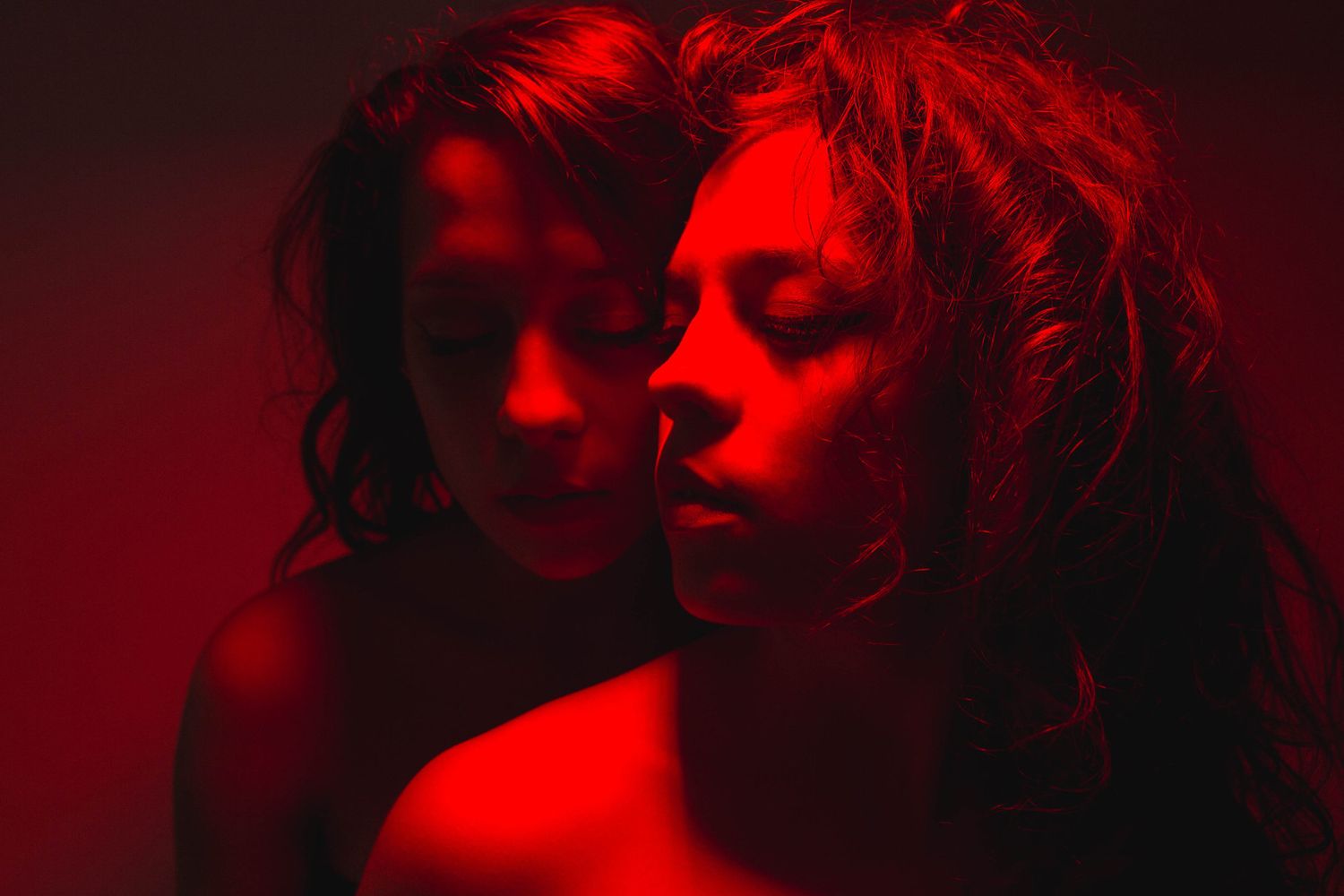 Two women in intimate pose bathed in moody red light with eyes closed against dark background.