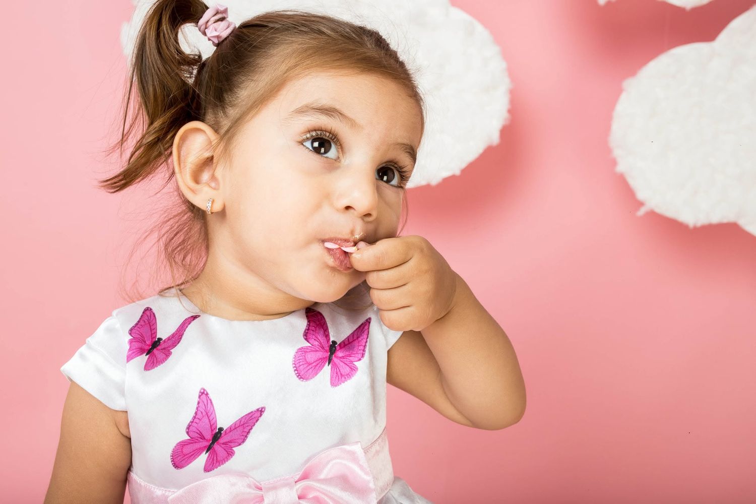 Adorable little girl in white butterfly dress with bunny ears poses thoughtfully against pink background.