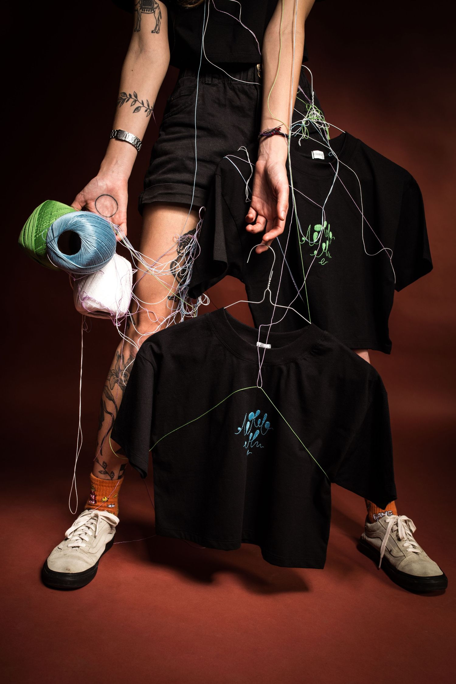 Person in black outfit holding colorful thread spools with tangled strings and black t-shirts on a dark red background.